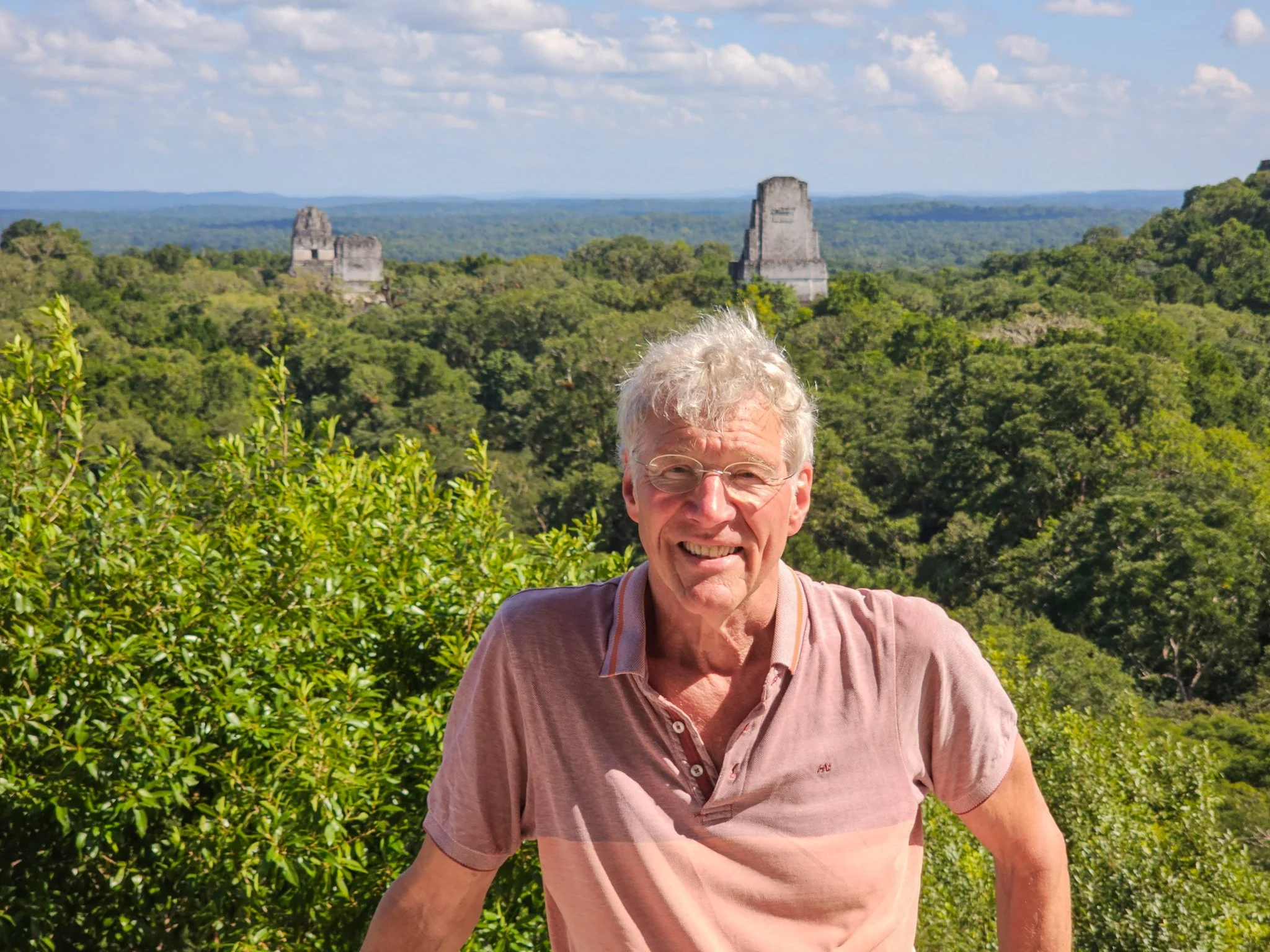A smiling man with gray curly hair and glasses stands in front of green jungle foliage with ancient Mayan ruins visible in the background under a blue sky.