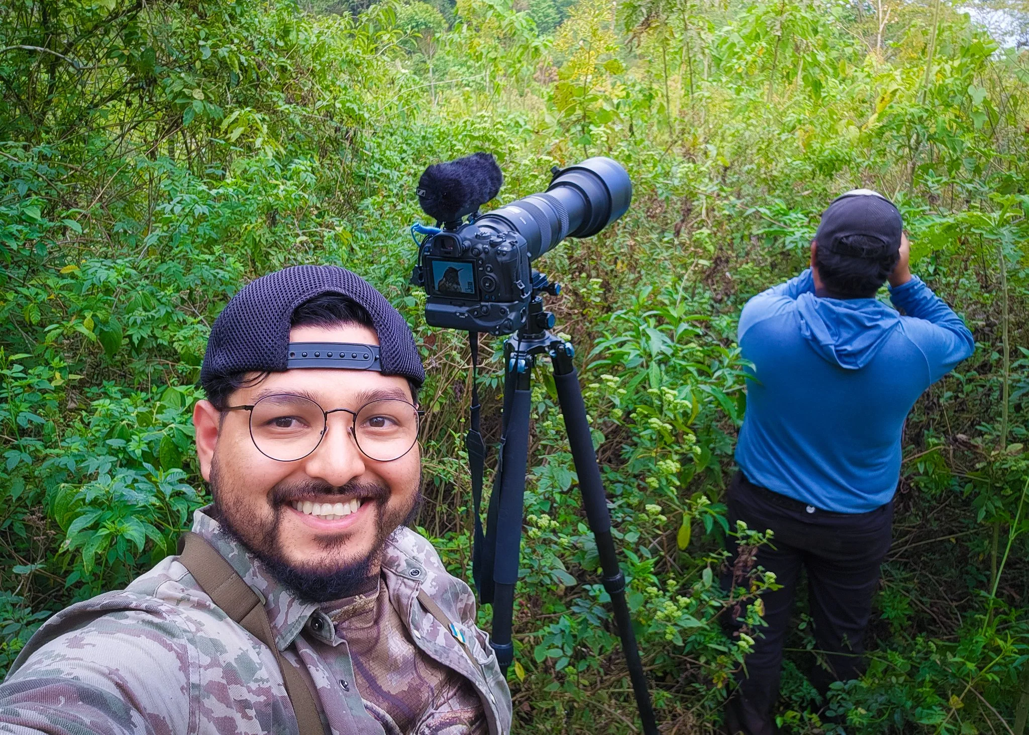 Two men in a lush green forest, one is taking a selfie while the other is using binoculars. There is a camera on a tripod pointed into the foliage, suggesting birdwatching or nature photography.