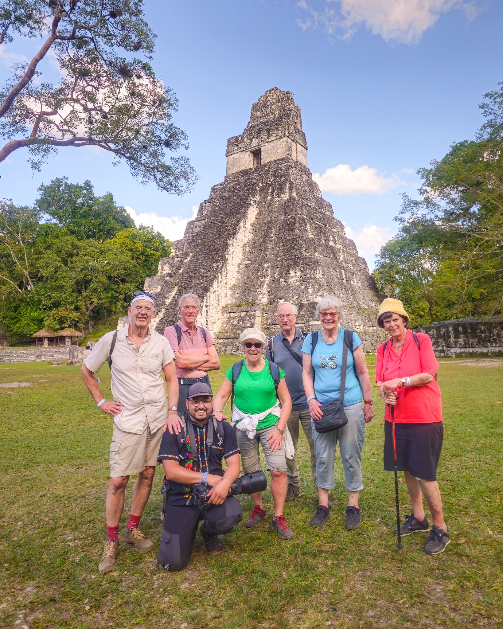 Group of people posing in front of a large Mayan pyramid at Tikal, surrounded by trees and grass.