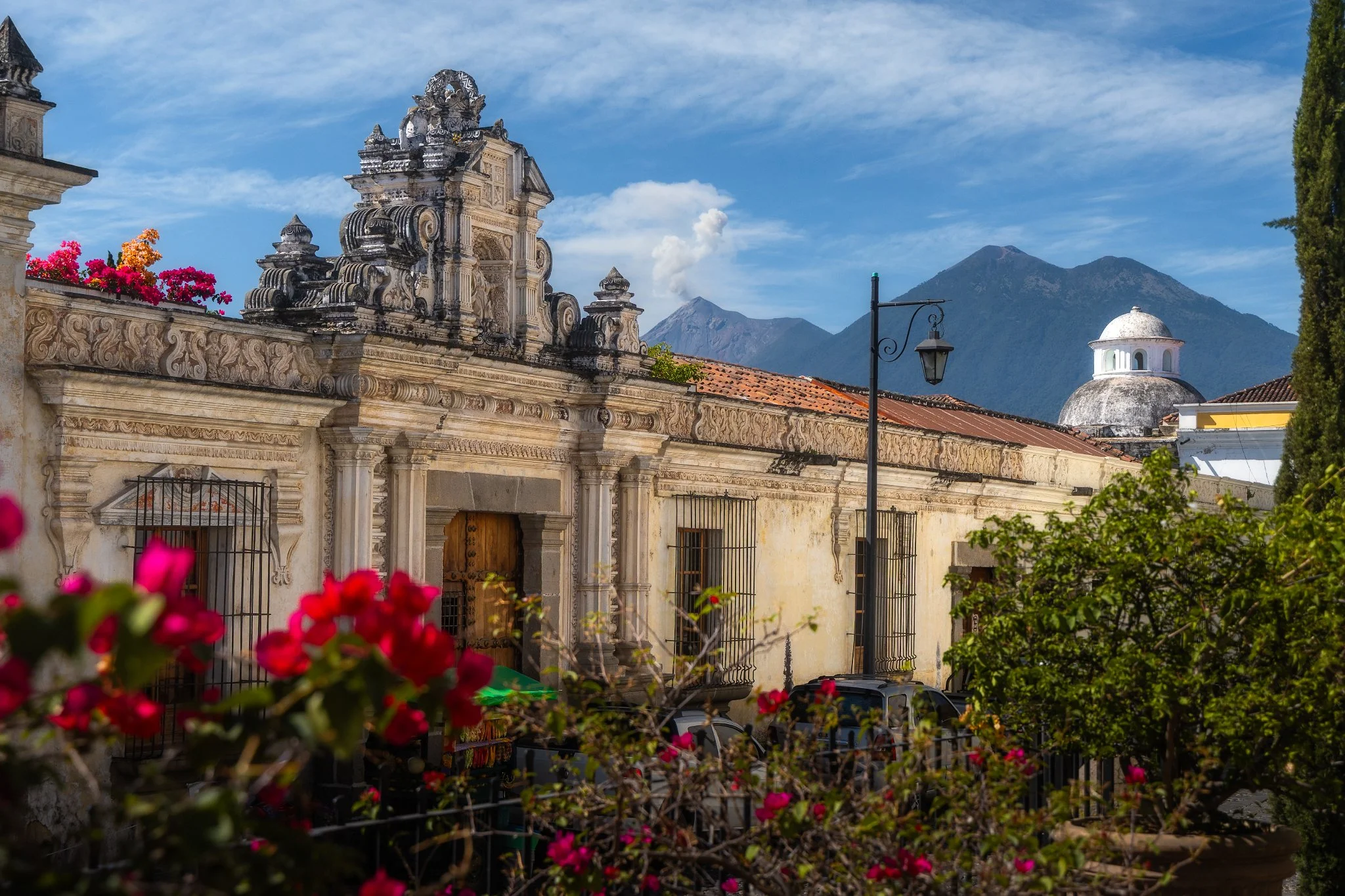 Historic stone building with ornate façade, flowering plants in the foreground, and mountains in the background under a partly cloudy sky.
