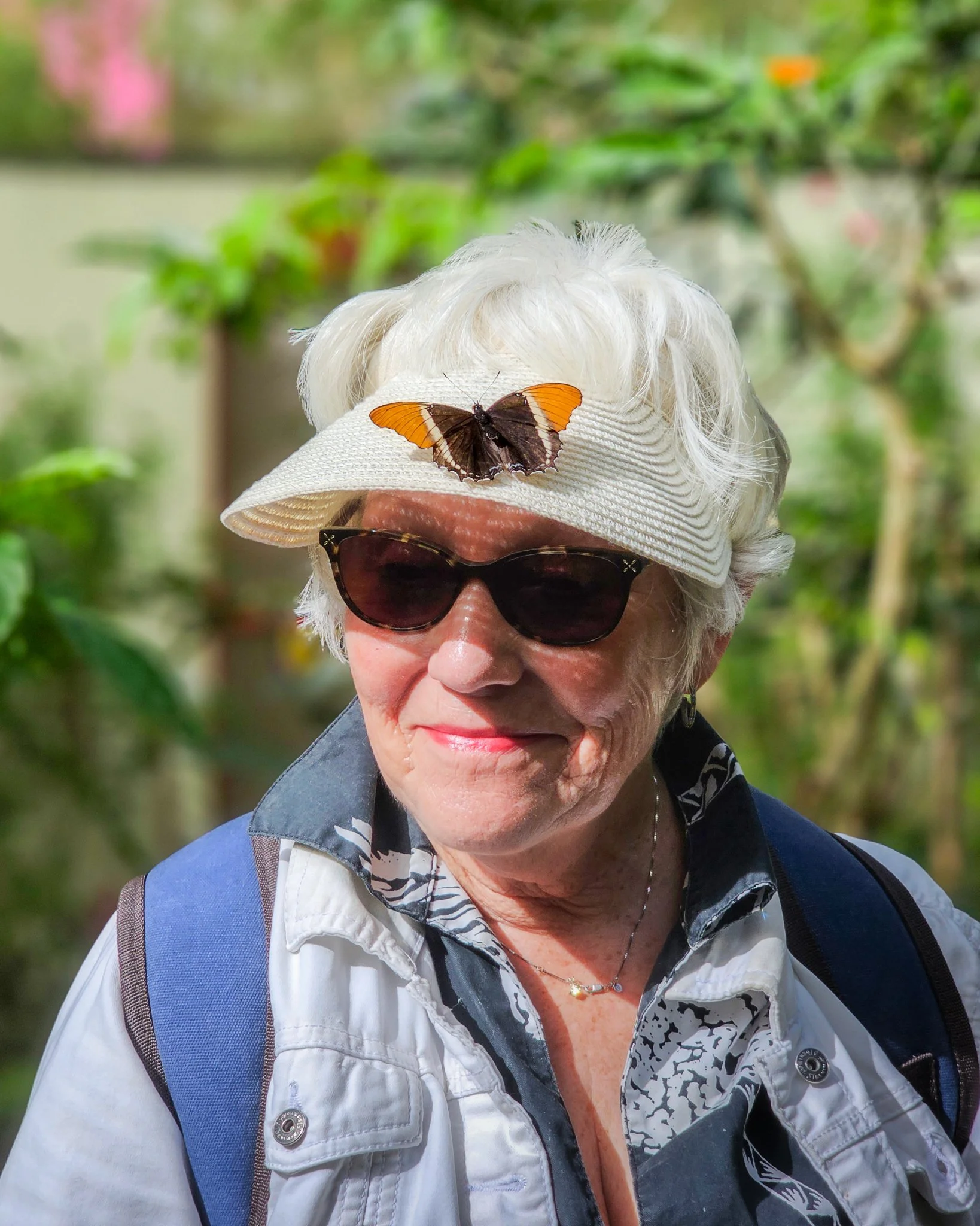 An elderly woman wearing sunglasses and a white hat with a butterfly on it, outdoors with green foliage in the background.