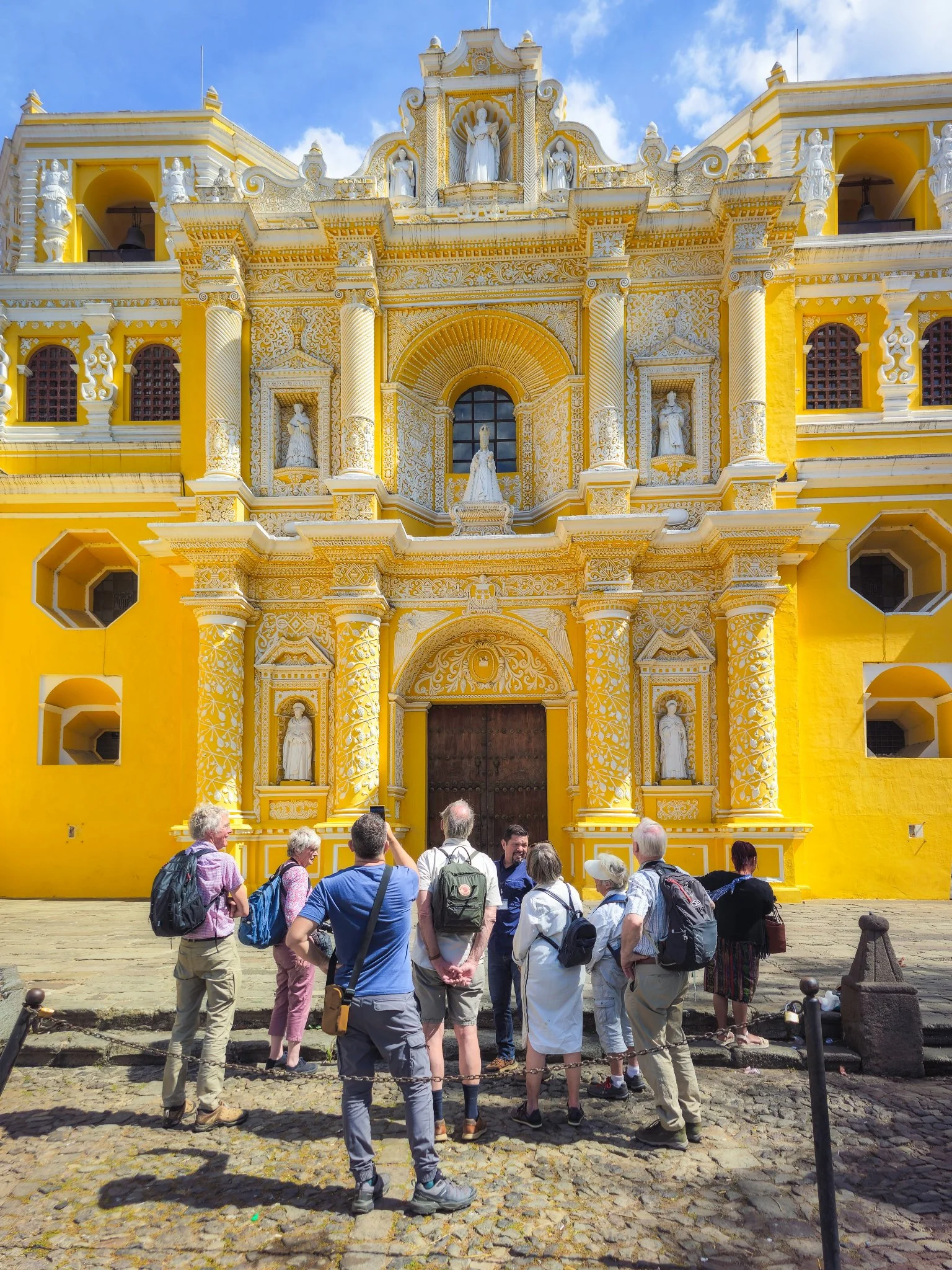 Tourists sightseeing in front of a yellow baroque-style church with elaborate white architectural details and statues.