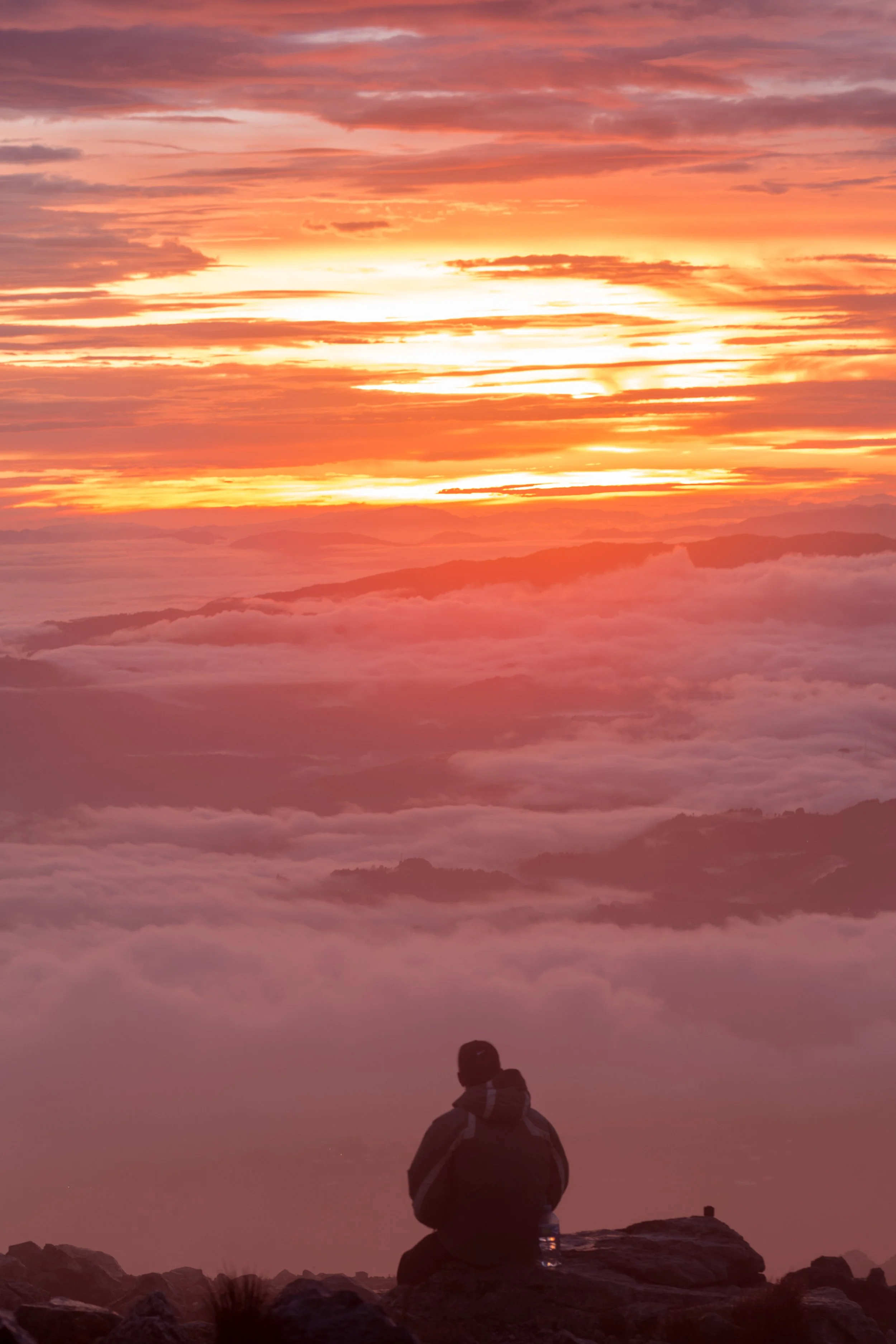 A person sitting on rocks watching a vibrant sunrise or sunset above clouds and distant mountains.
