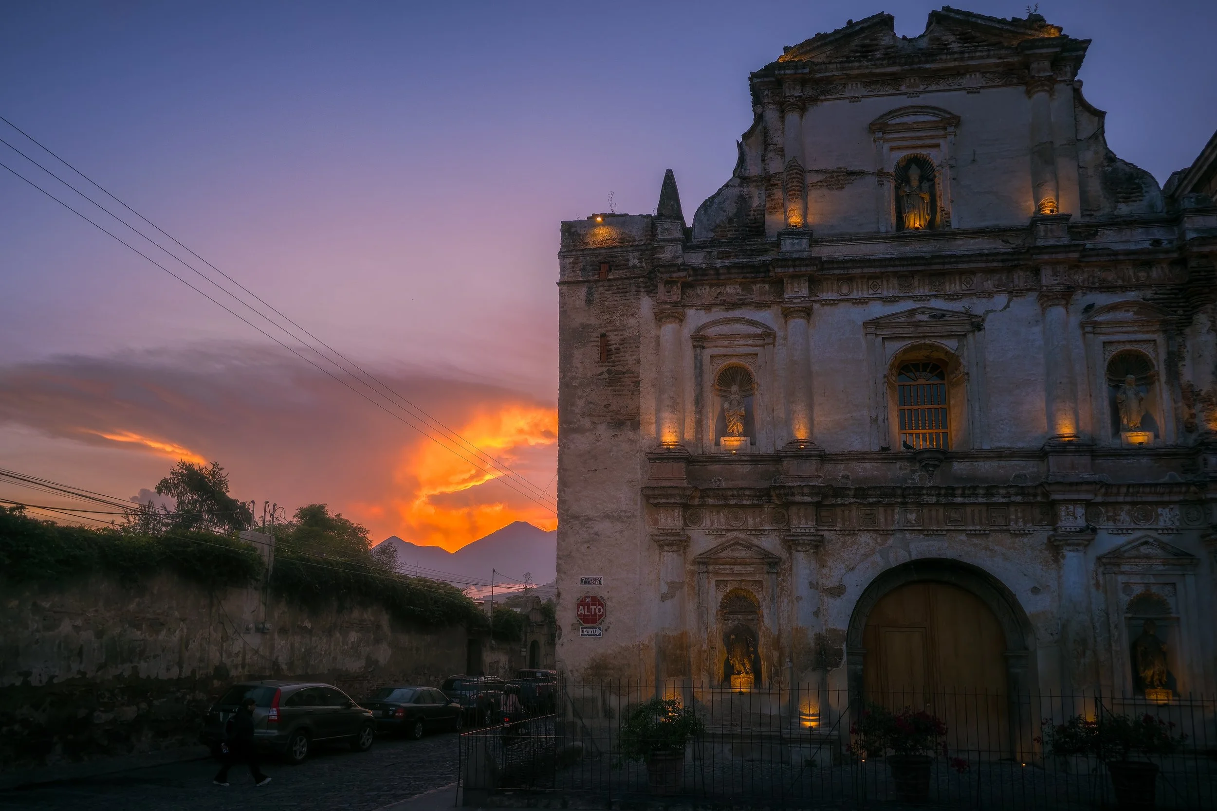 Sunset behind an old, illuminated church with statues, overlooking parked cars and a cobblestone street.