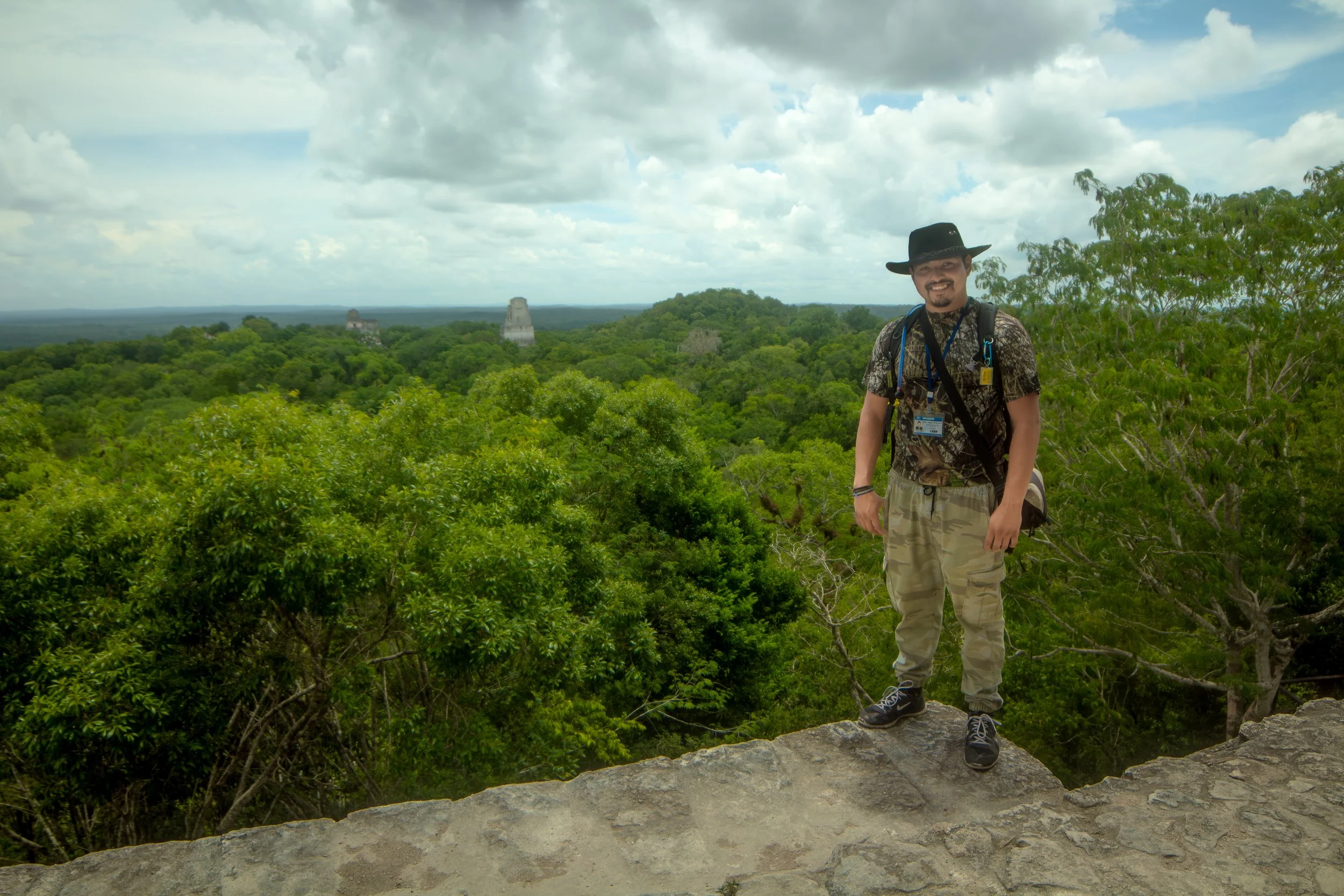 A man wearing a black hat, camo pants, and a patterned shirt standing on a stone ledge amidst lush green trees with Mayan ruins visible in the distance under a cloudy sky.