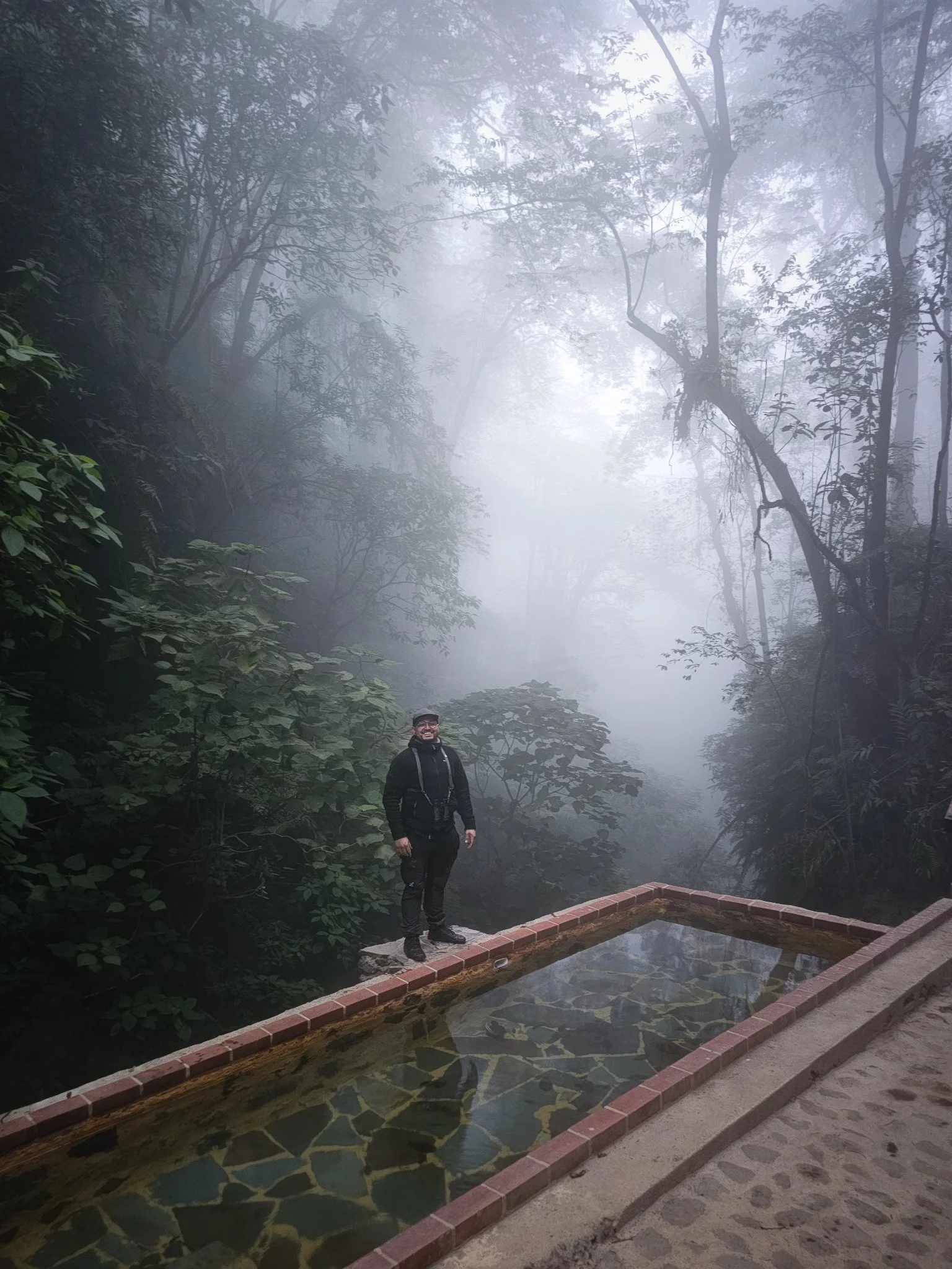 A person standing near a small water feature with a foggy forest in the background.