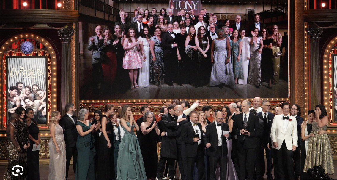 A group of people dressed in formal attire on stage at an awards ceremony, with a large screen behind them displaying a group photo from a previous event.  Join a fellow group of investors supporting commercial-stage and screen narratives.