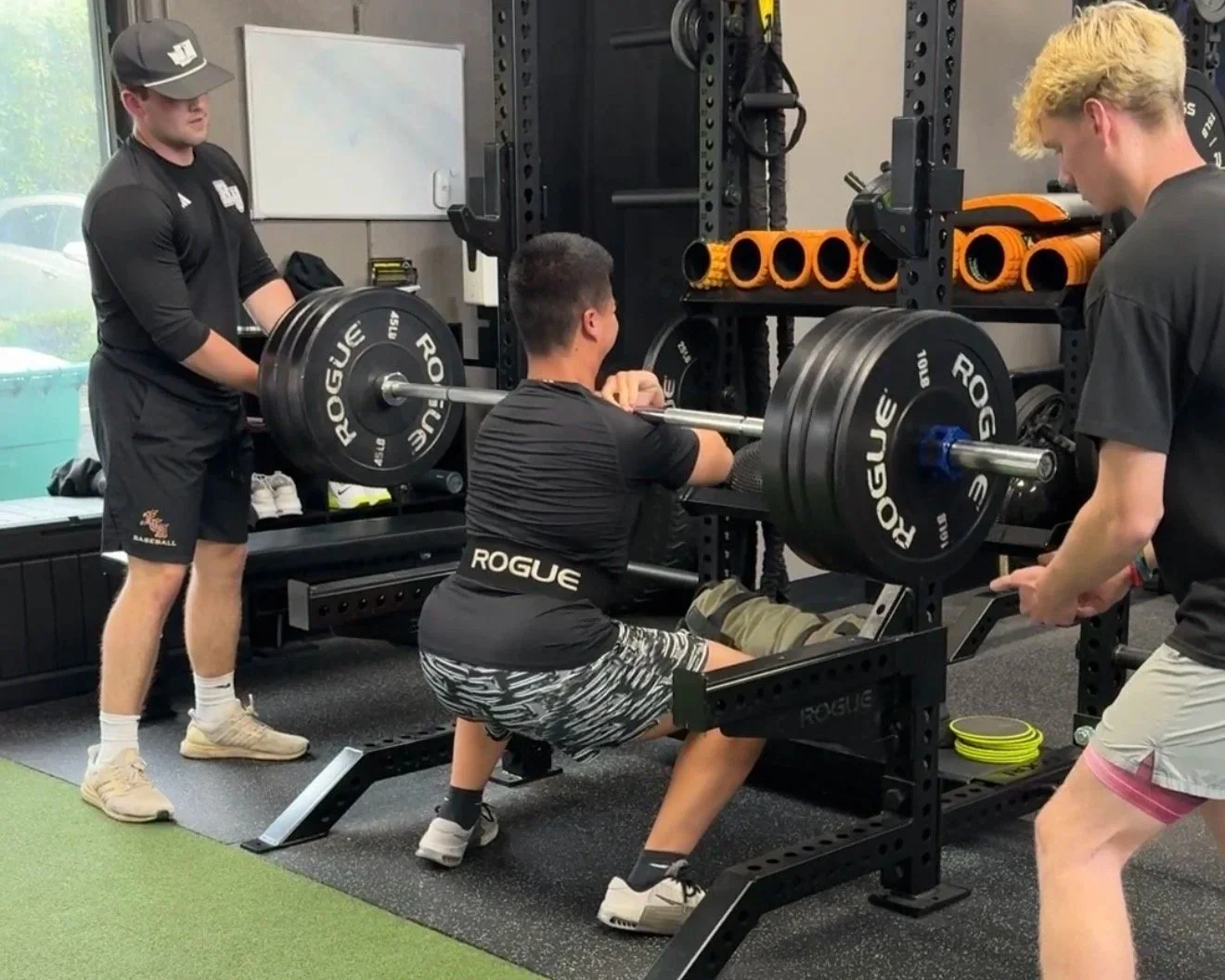 A young man performs a squat with a loaded barbell on a squat rack while two men assist and spot him during a workout in a gym.