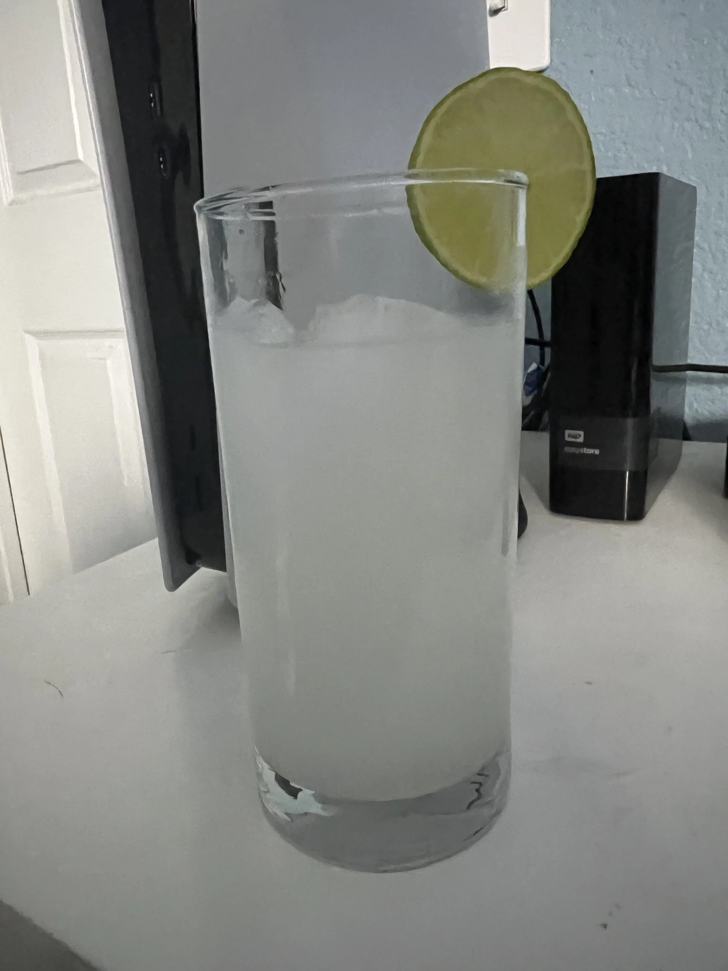 A tall glass with a slice of lime on the rim, filled with a clear beverage, placed on a white surface.