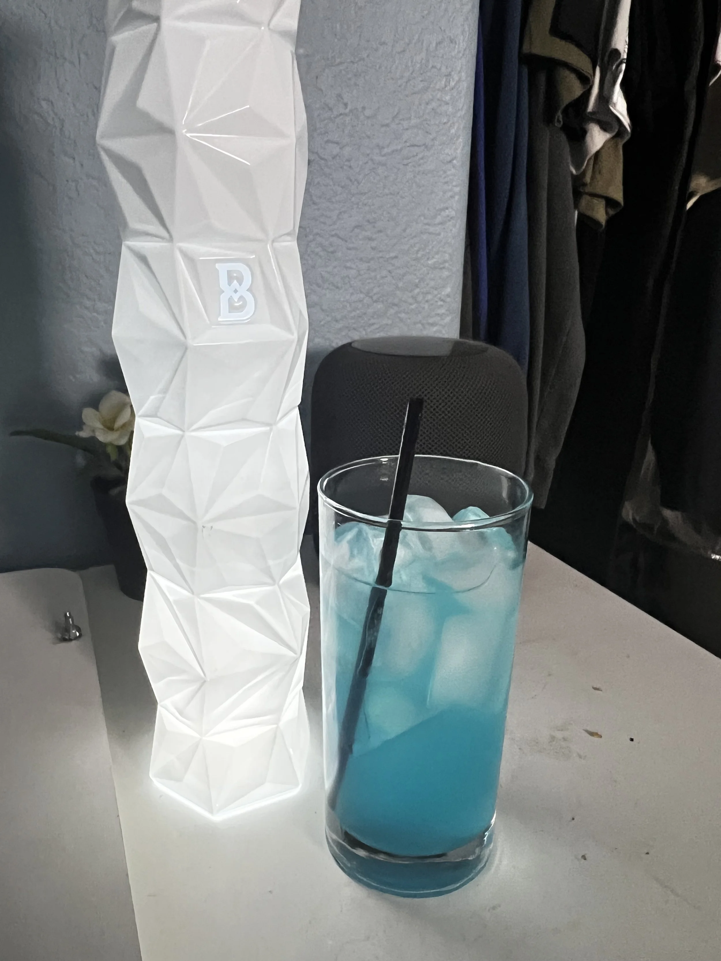 A tall glass with blue liquid and ice, with a black straw, placed on a white table.