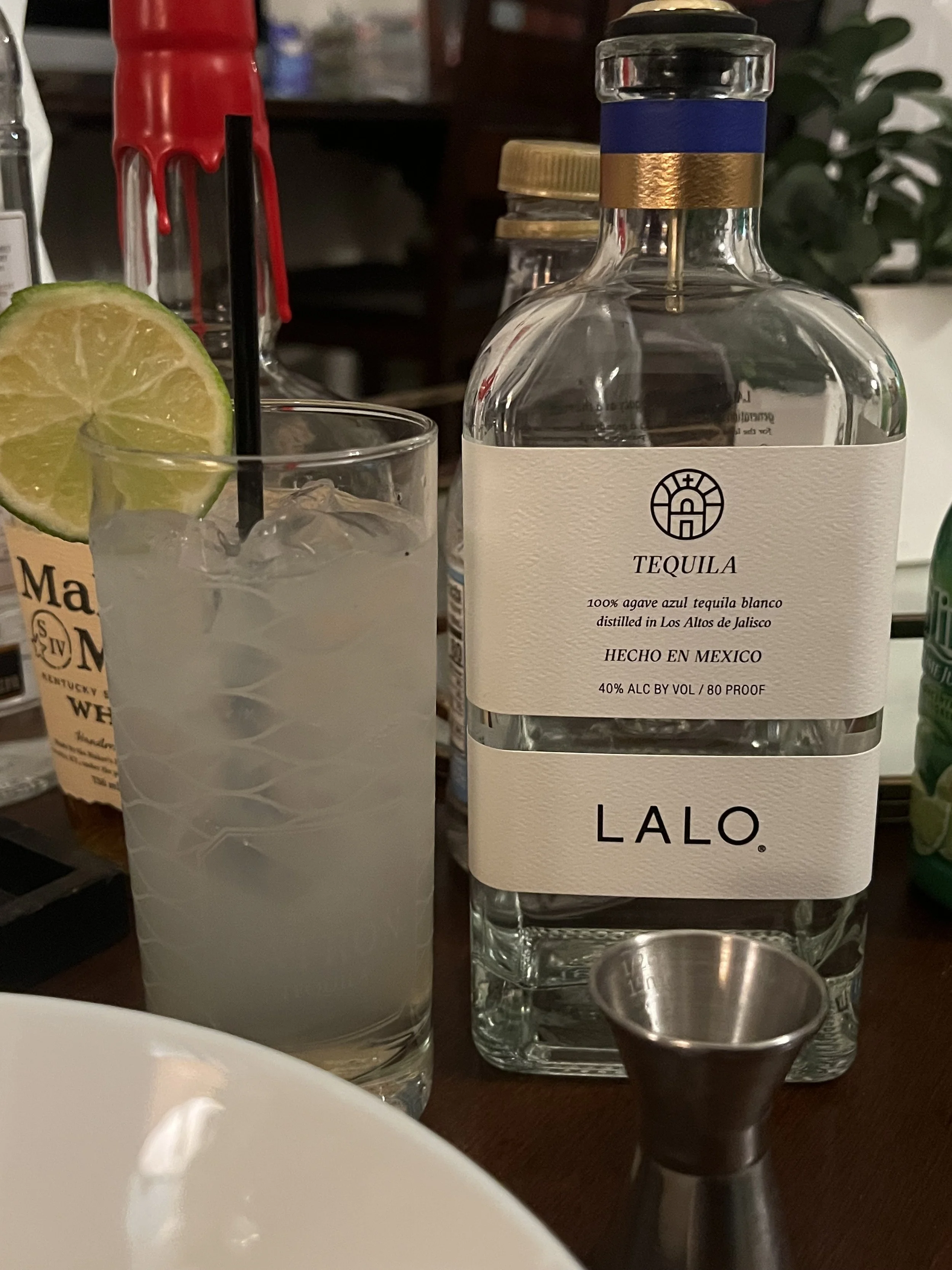 A glass of margarita with a lime slice and a straw, a bottle of LALO tequila, and a metal jigger on a bar counter.