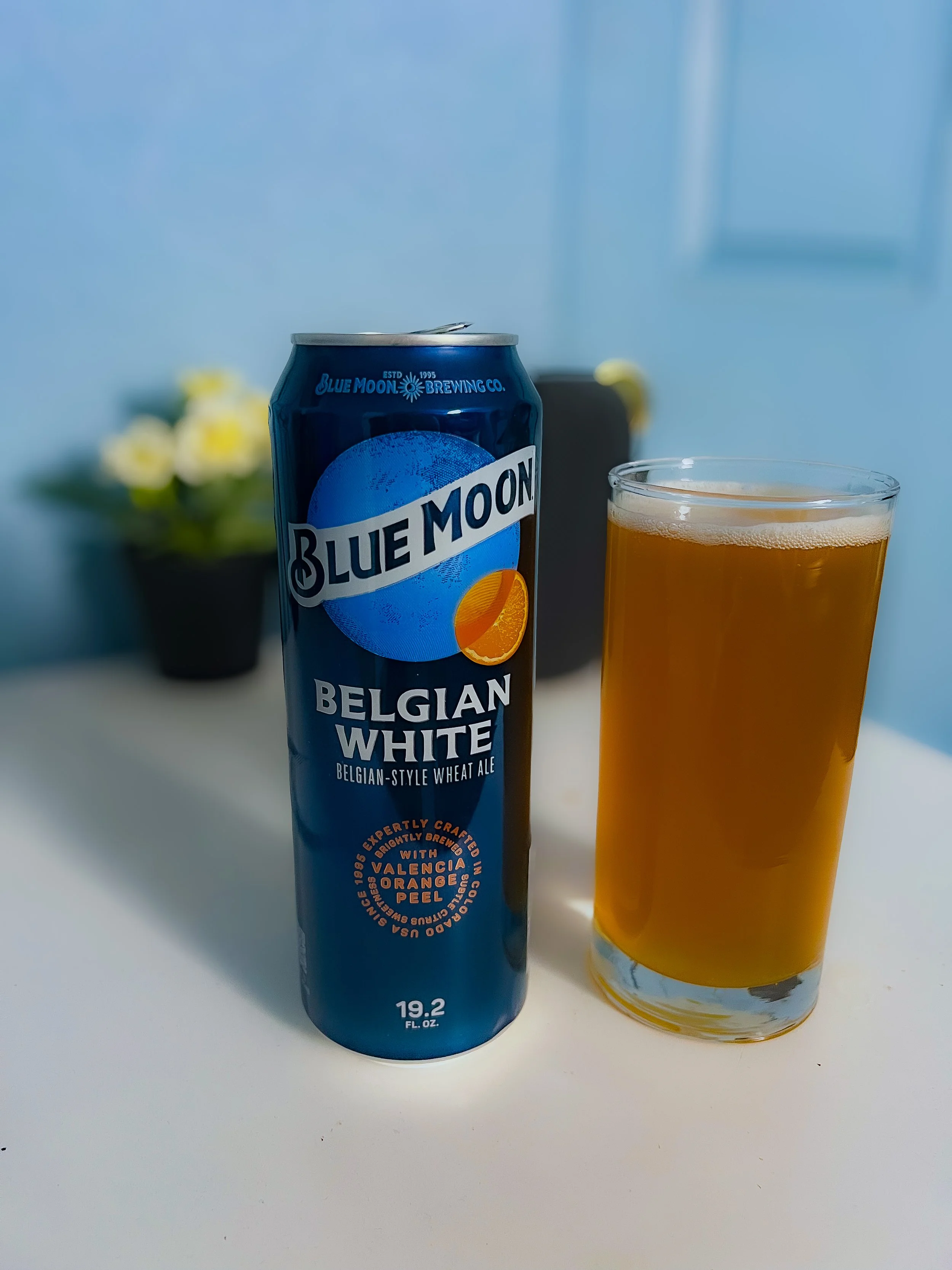 A can of Blue Moon Belgian White beer next to a glass of beer with a foamy head on a white surface. In the background, a blurred black pot with yellow and white flowers on a blue wall.