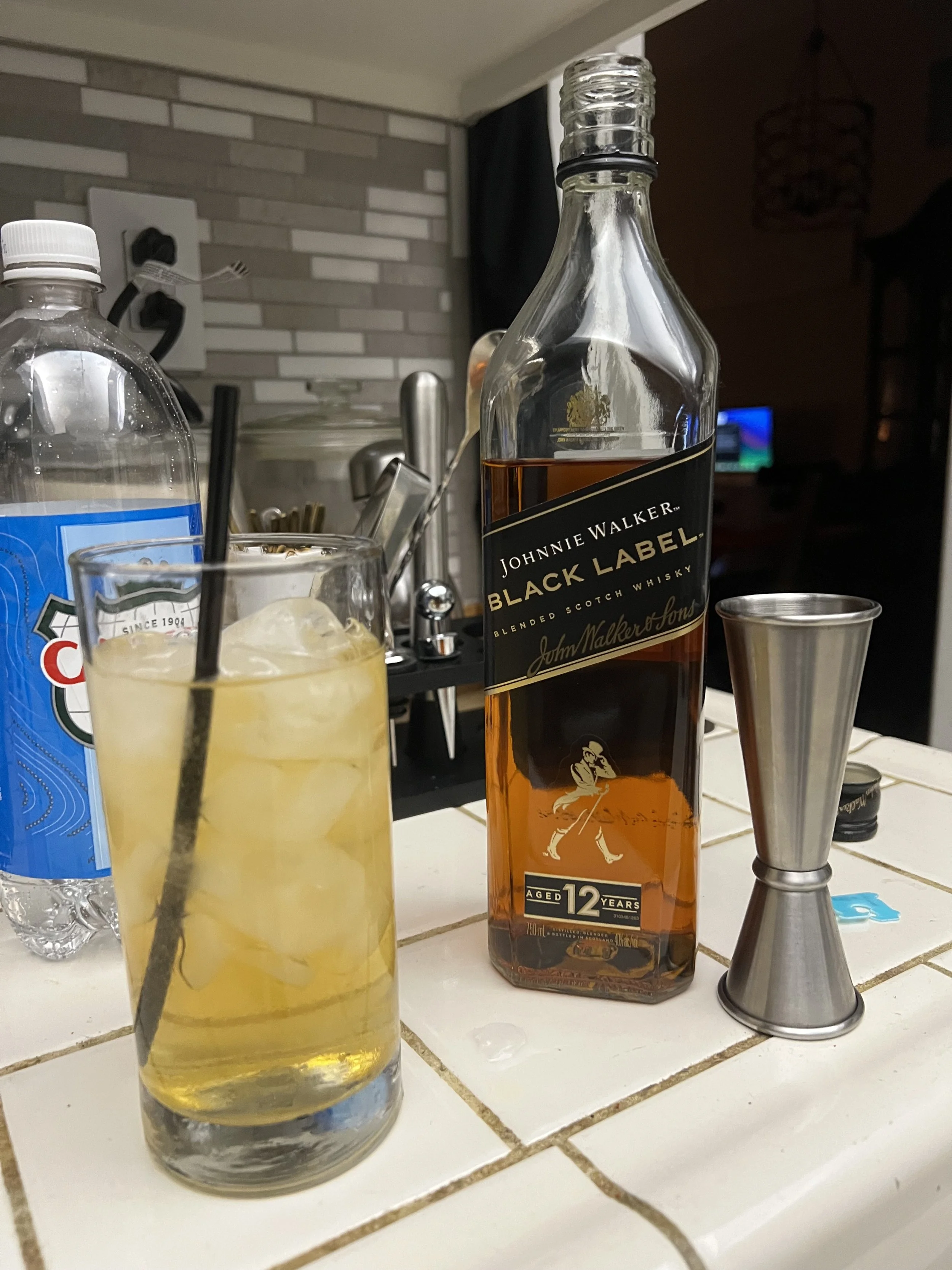 A Highball Cocktail with ice and a black straw, a bottle of Johnnie Walker Black Label whiskey, a metal jigger, and a bottle of soda water on a kitchen counter.