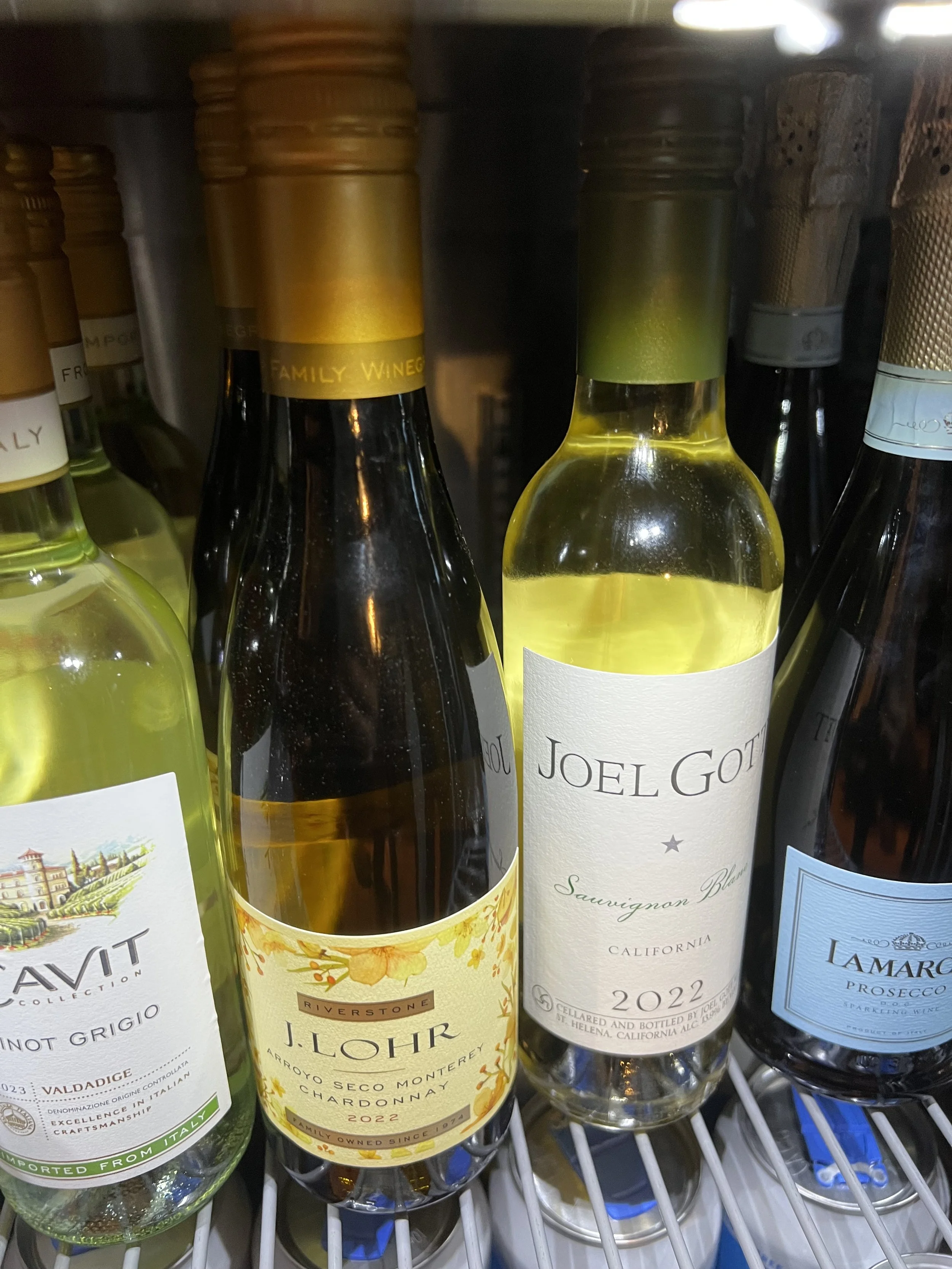 Selection of wine bottles on a refrigerated shelf, including Sauvignon Blanc by Joel Gott, J. Lohr Chardonnay, and others.