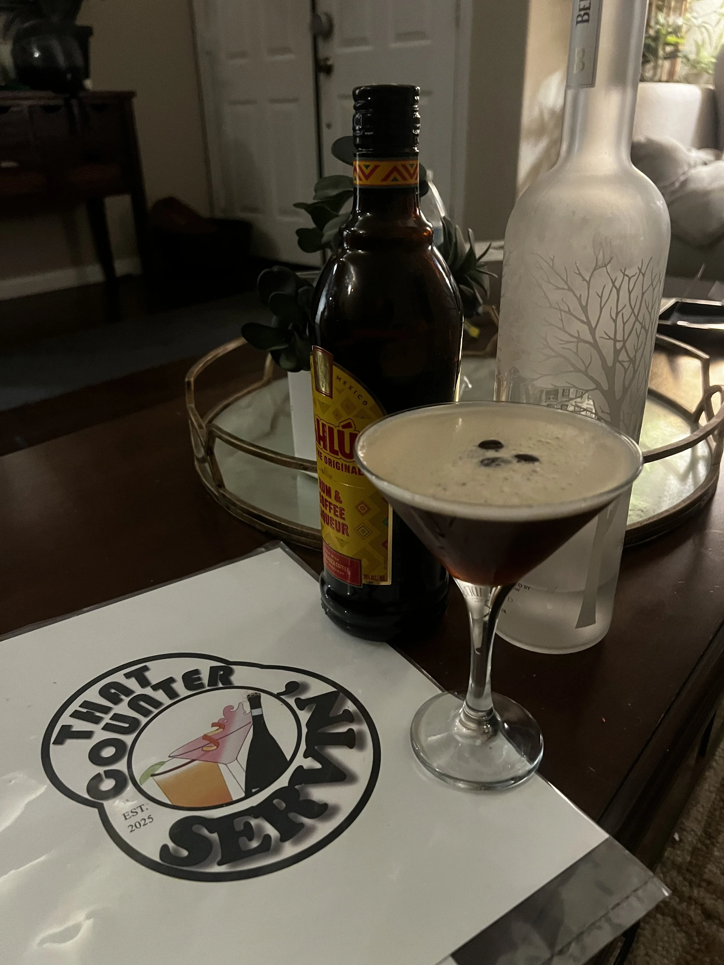 A cocktail in a martini glass with a frothy top and dark liquid underneath on a table, next to a bottle of Coffee Liquer,a Bottle of Belvedere Vodka and a tray with greenery in a cozy indoor setting.