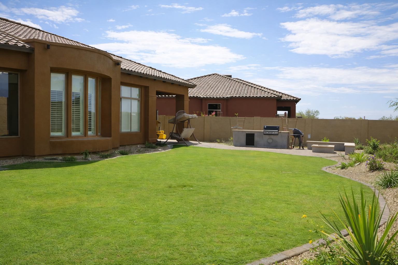 Backyard of a house with a well-maintained green lawn, outdoor seating, a barbecue grill, and a worker near the fence under a partly cloudy sky.