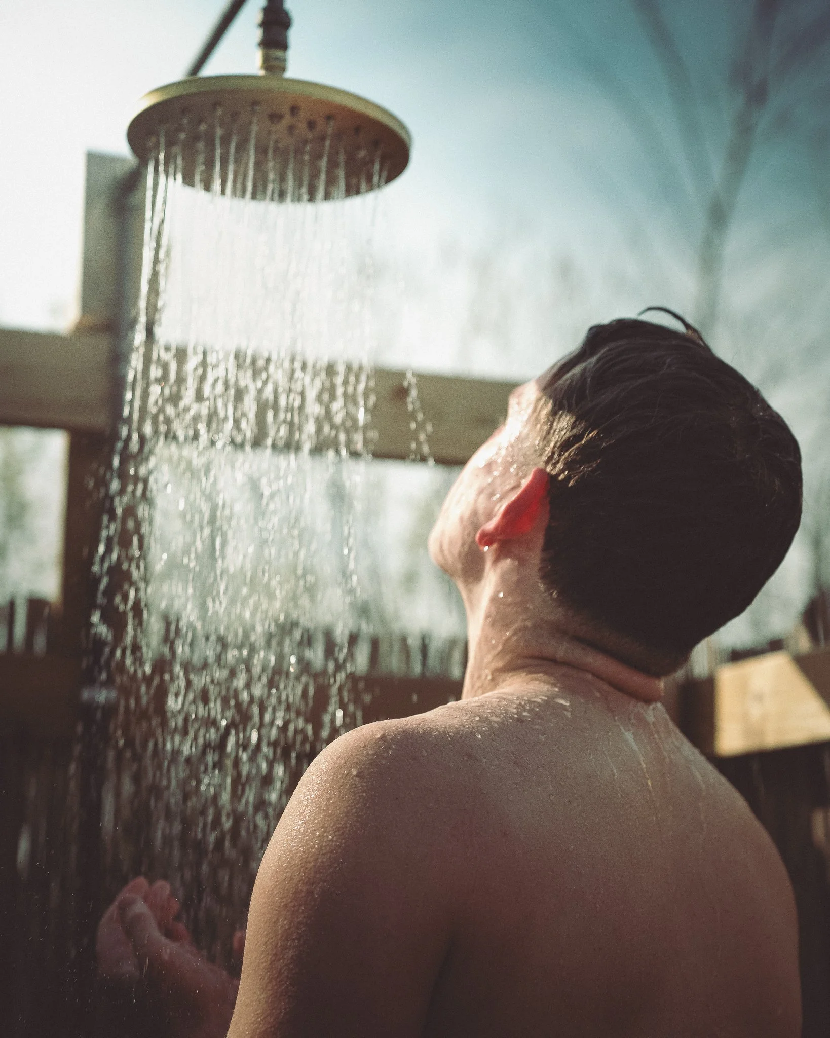 At Unfold Sauna Club a Person with wet hair under an outdoor shower with water spraying from the showerhead, back view.