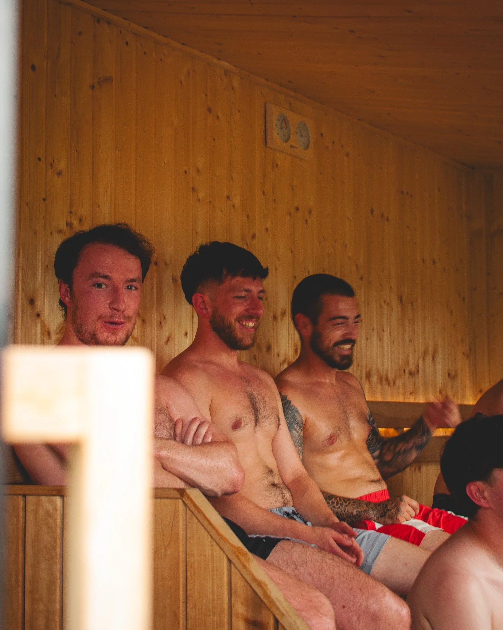 At Unfold Sauna Club Three men sitting shirtless in a sauna with wood paneling, smiling and talking.