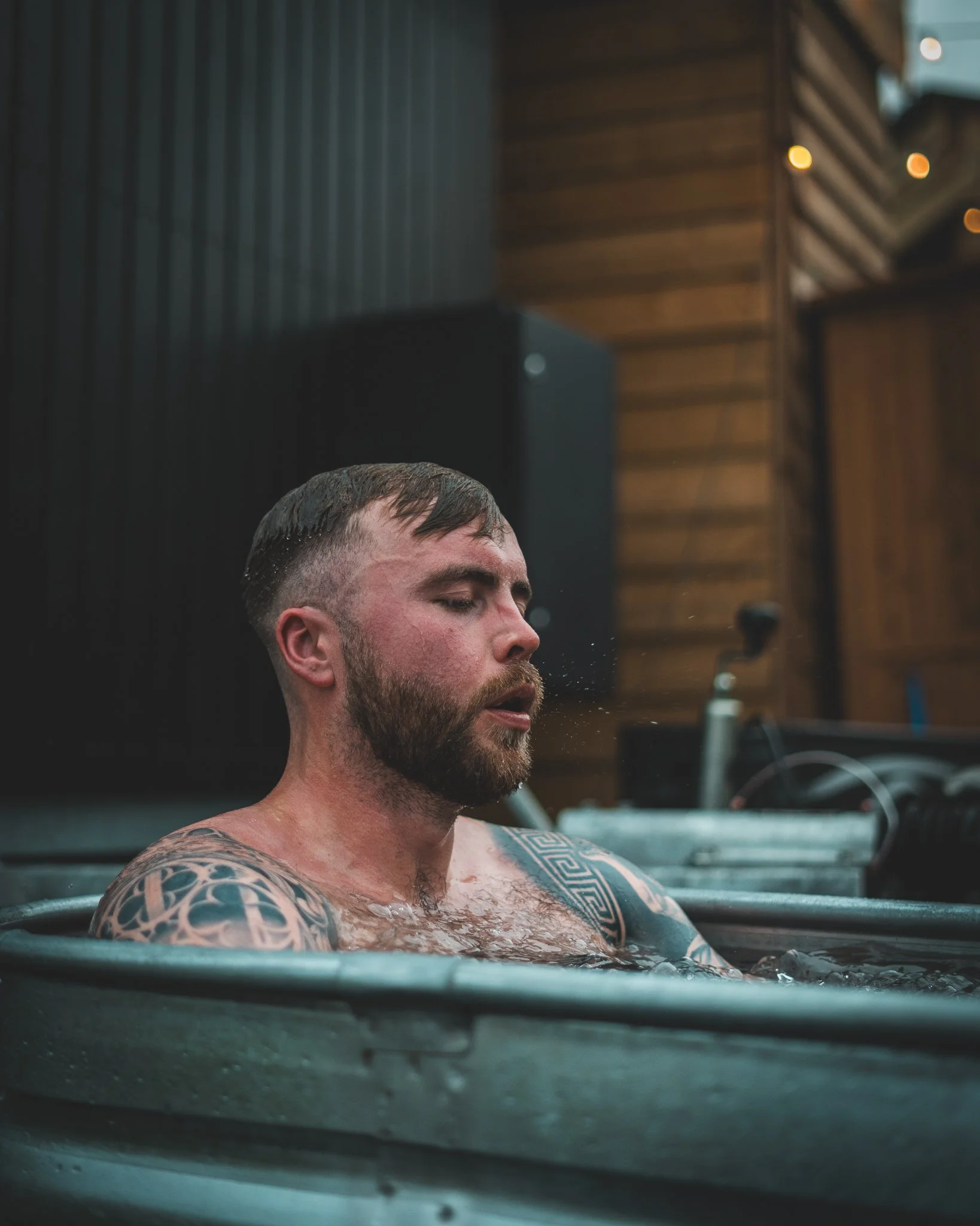 At Unfold Sauna Club A man with tattoos on his arms and beard, soaking in a hot tub, with his eyes closed and water splashing against his face.