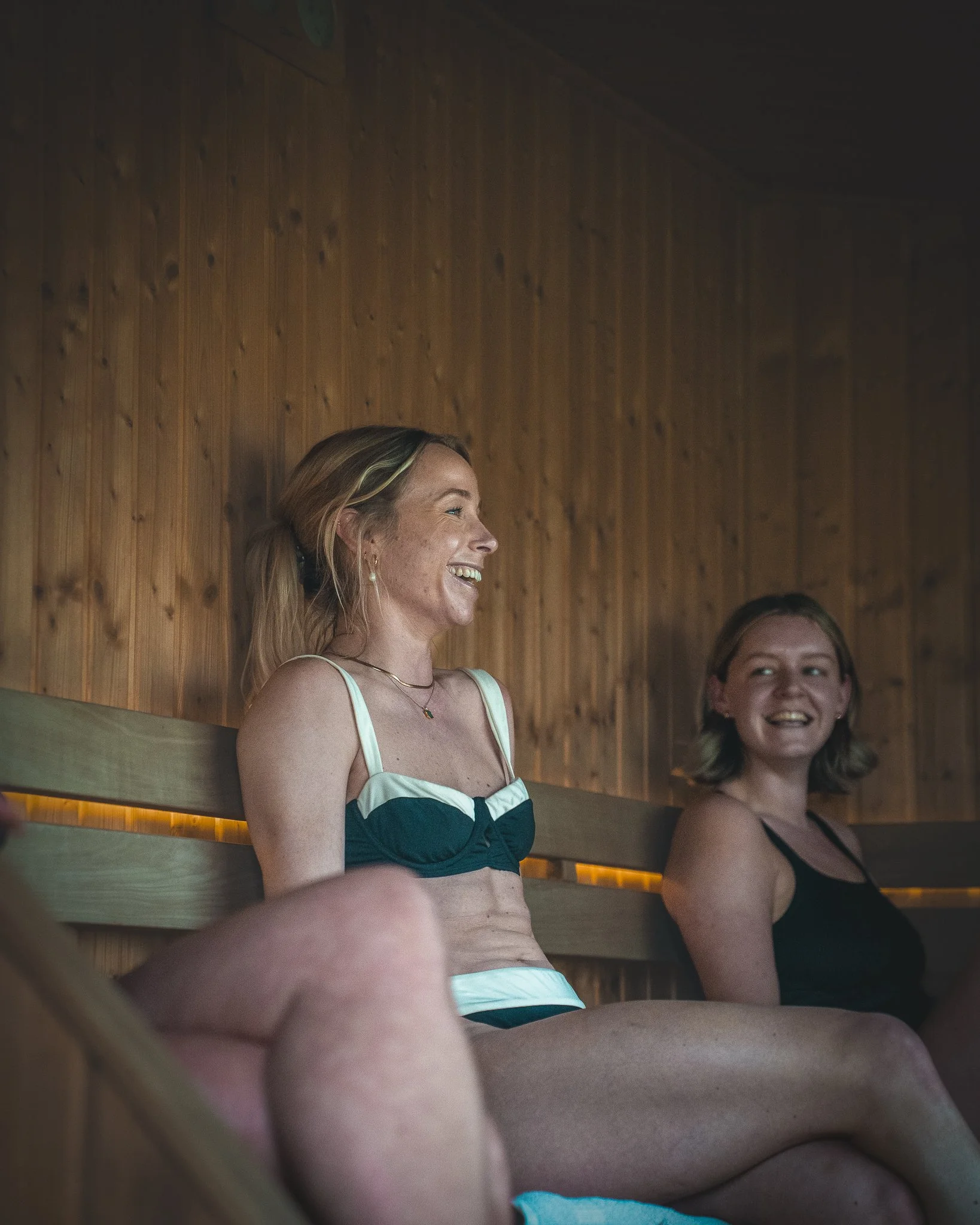At Unfold Sauna Club Two women sitting in a sauna, smiling and enjoying conversation, with wood-paneled walls in the background.