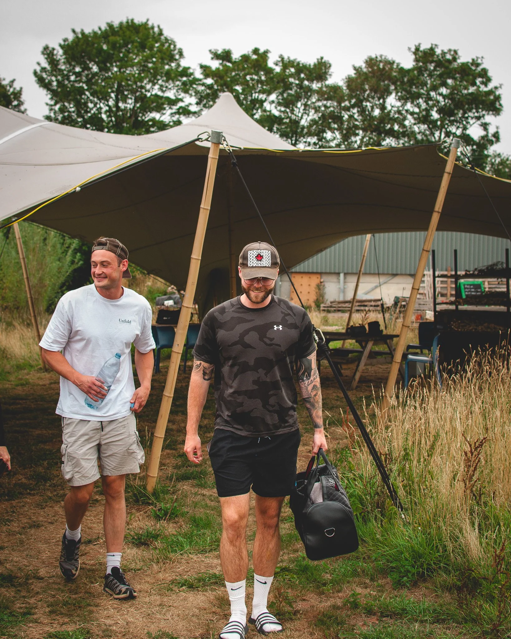At Unfold Sauna Club Two men walking outdoors near a white canopy tent, one holding a water bottle and the other carrying a sports bag, in a grassy area with trees in the background.