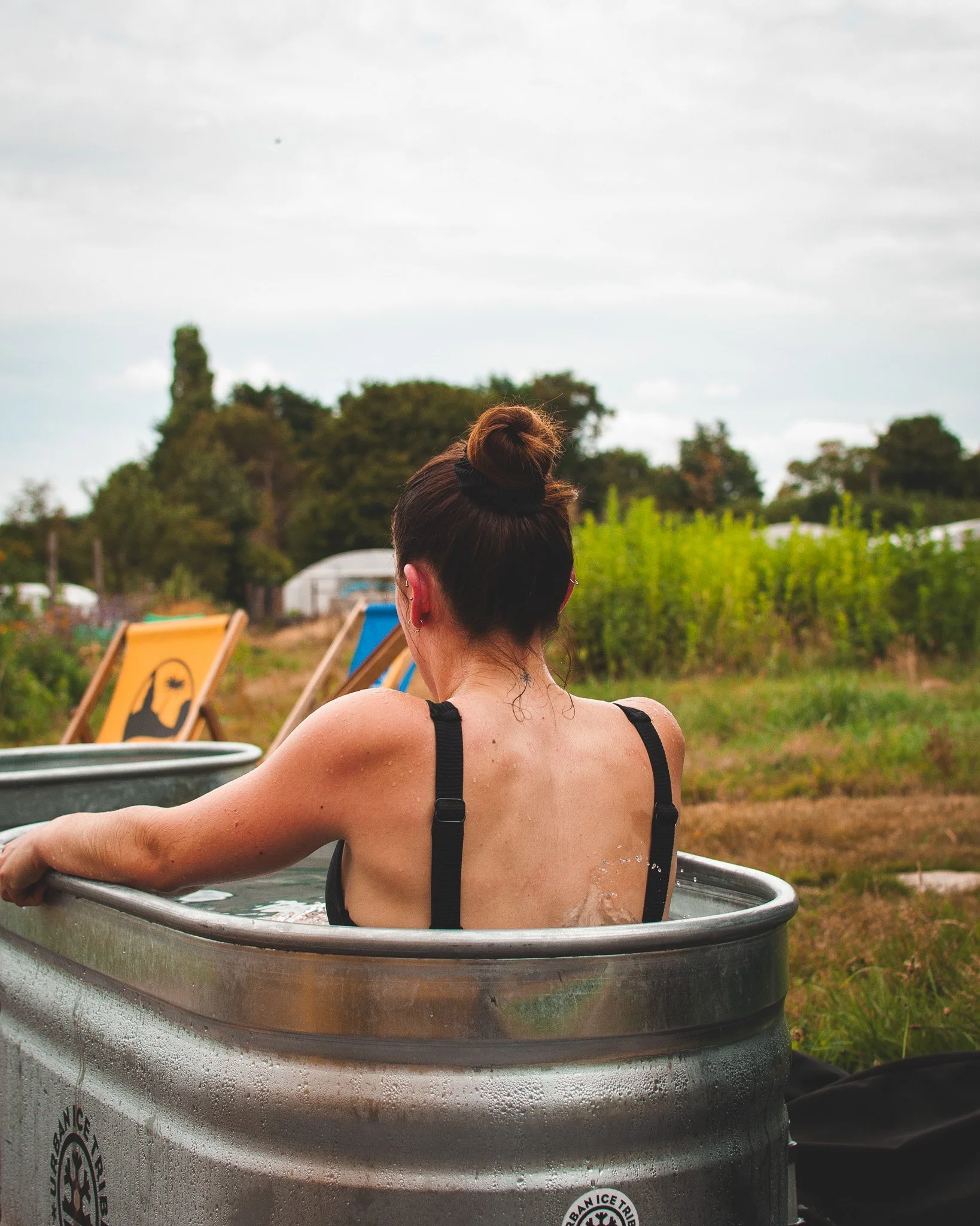 At Unfold Sauna Club A woman with a bun hairstyle, wearing a black bra, sitting in a metal tub outdoors, with greenery and lounge chairs in the background.