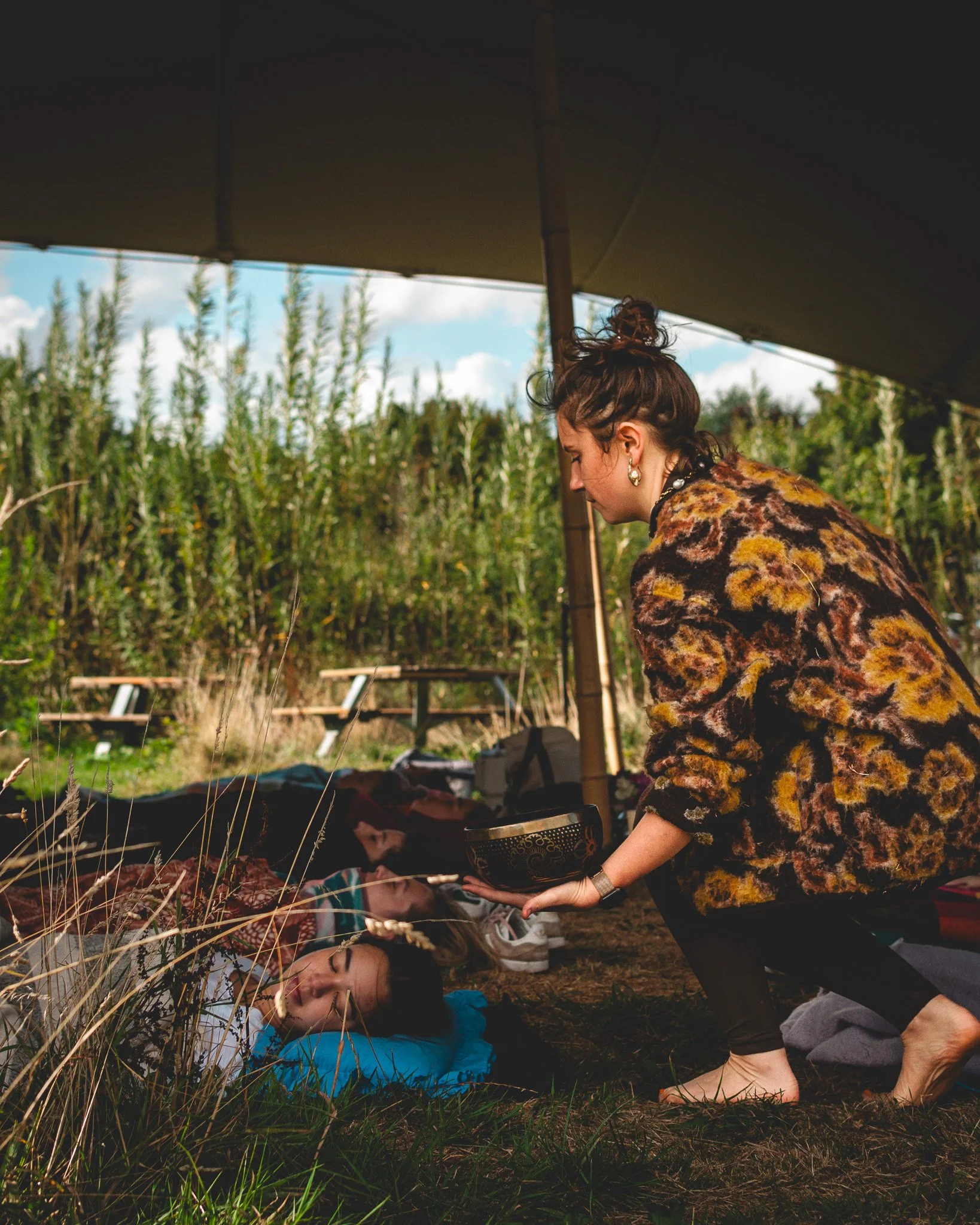 A woman is performing a spiritual or healing ritual on people lying under a tent outdoors in a grassy area with tall plants in the background At Unfold Sauna Club.