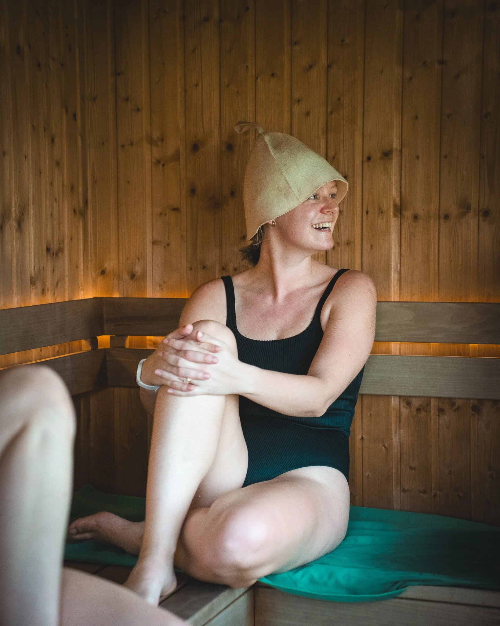 At Unfold Sauna Club A woman sitting cross-legged in a wooden sauna, wearing a black swimsuit and a felt hat, smiling and looking to her right.