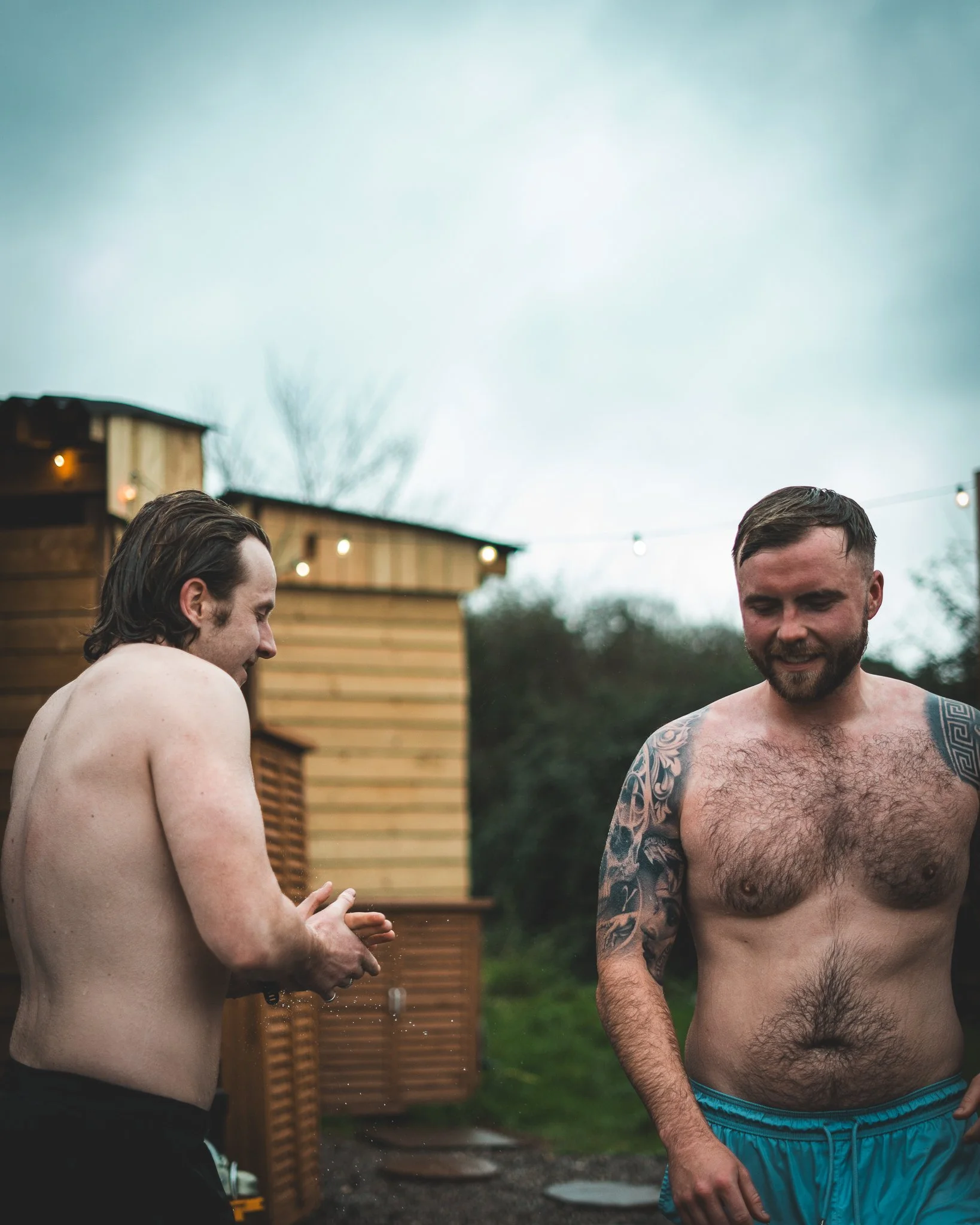 At Unfold Sauna Club Two shirtless men standing outside near a wooden shed, smiling and talking, with string lights hanging overhead and a cloudy sky in the background.
