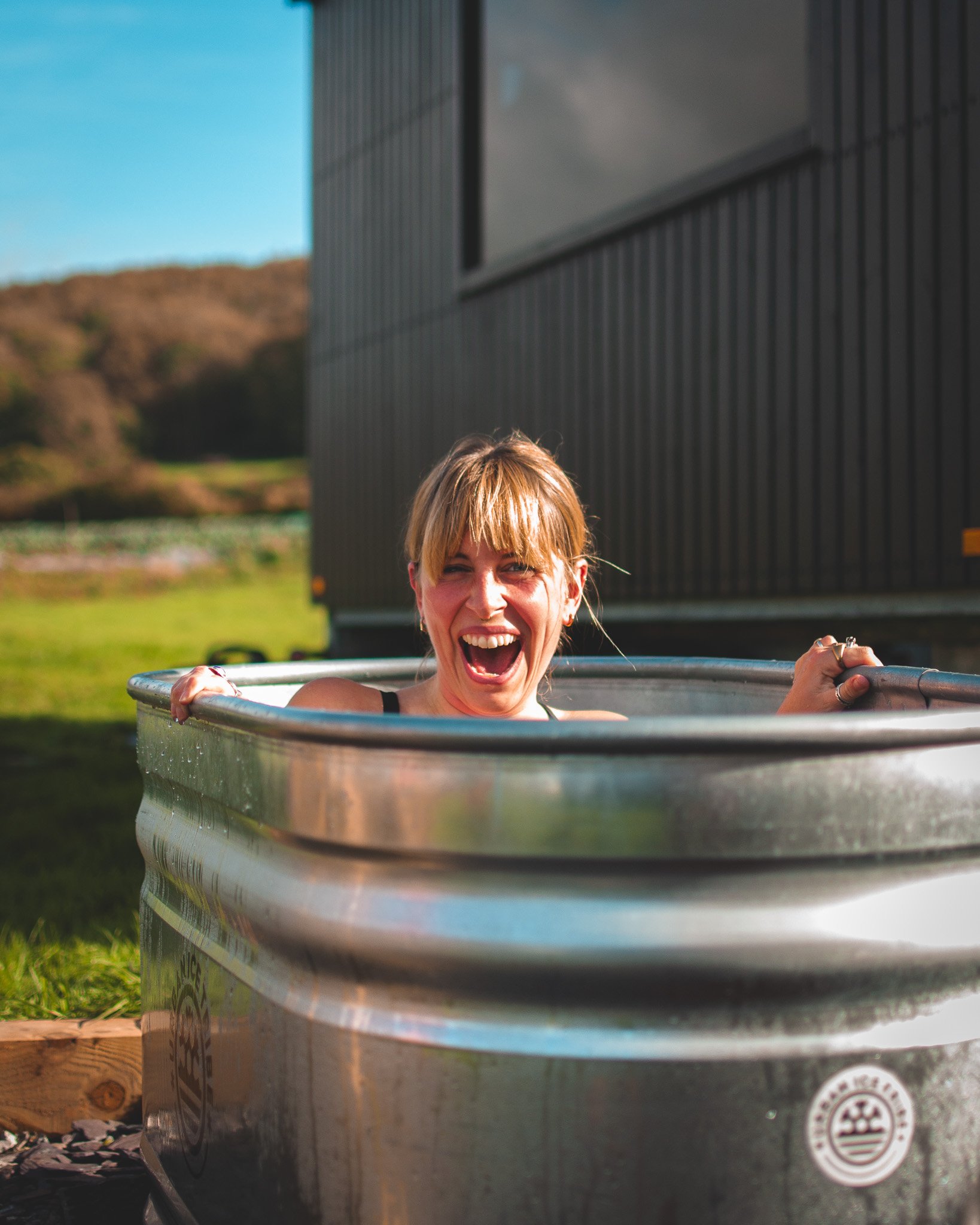 At Unfold Sauna Club Woman laughing and smiling while sitting in a metal tub outdoors on grass, with a building and hills in the background.