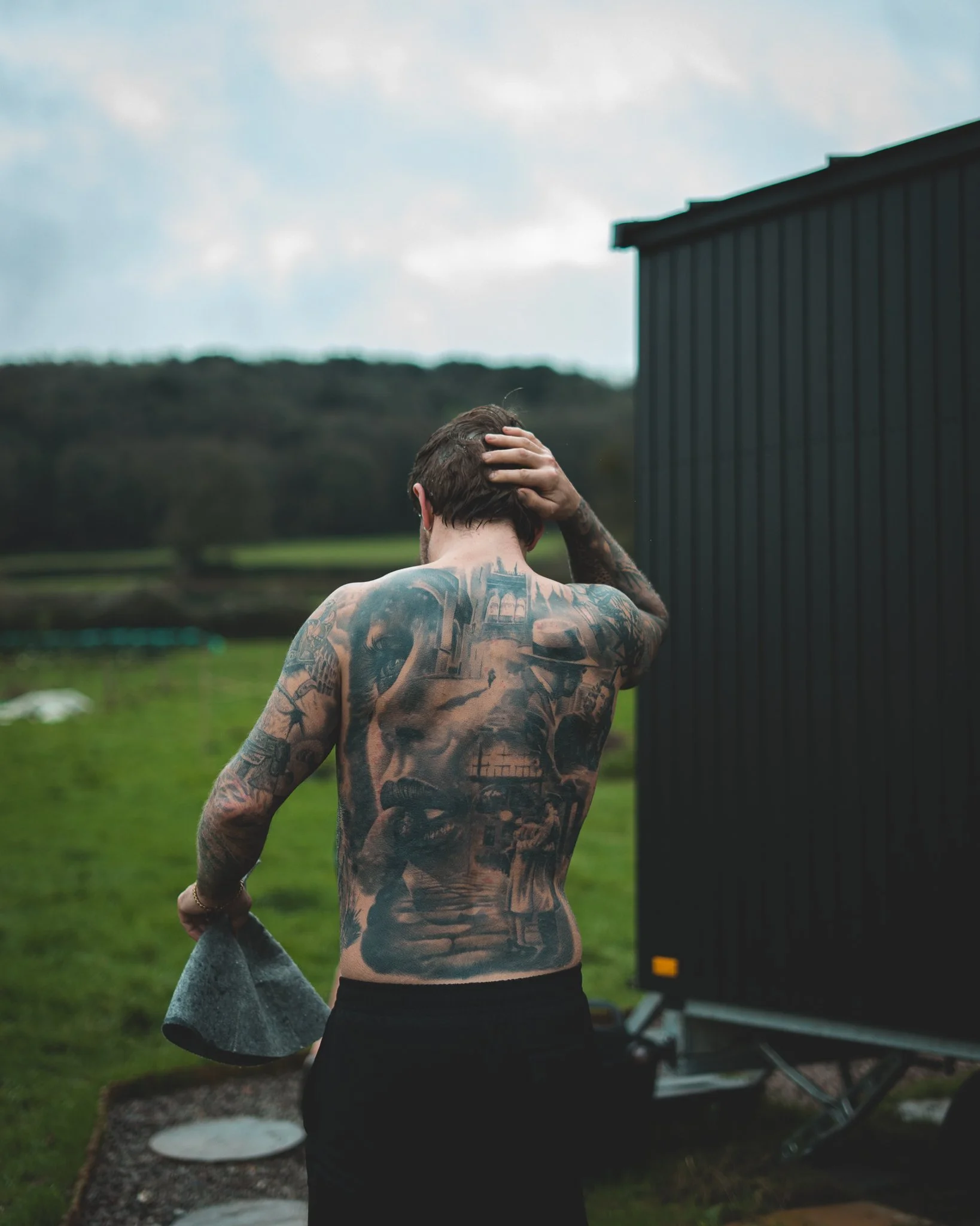 At Unfold Sauna Club A shirtless man with tattoos on his back and arms standing outdoors with his hand on his head, near a black trailer in a grassy field.