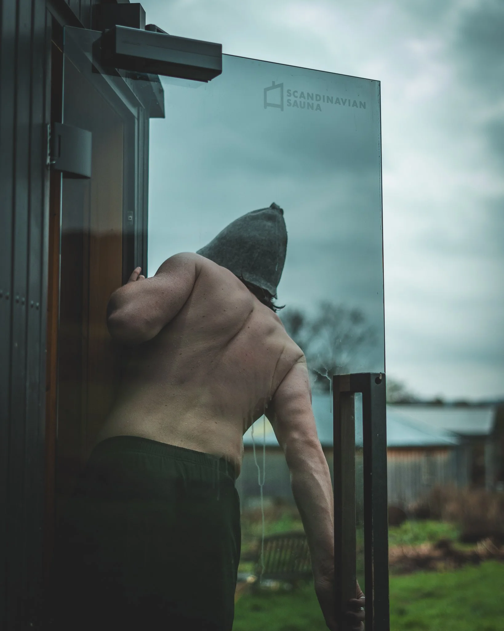 At Unfold Sauna Club A shirtless man with a beanie on his head opens a glass door marked 'Scandinavian Sauna' and looks inside.