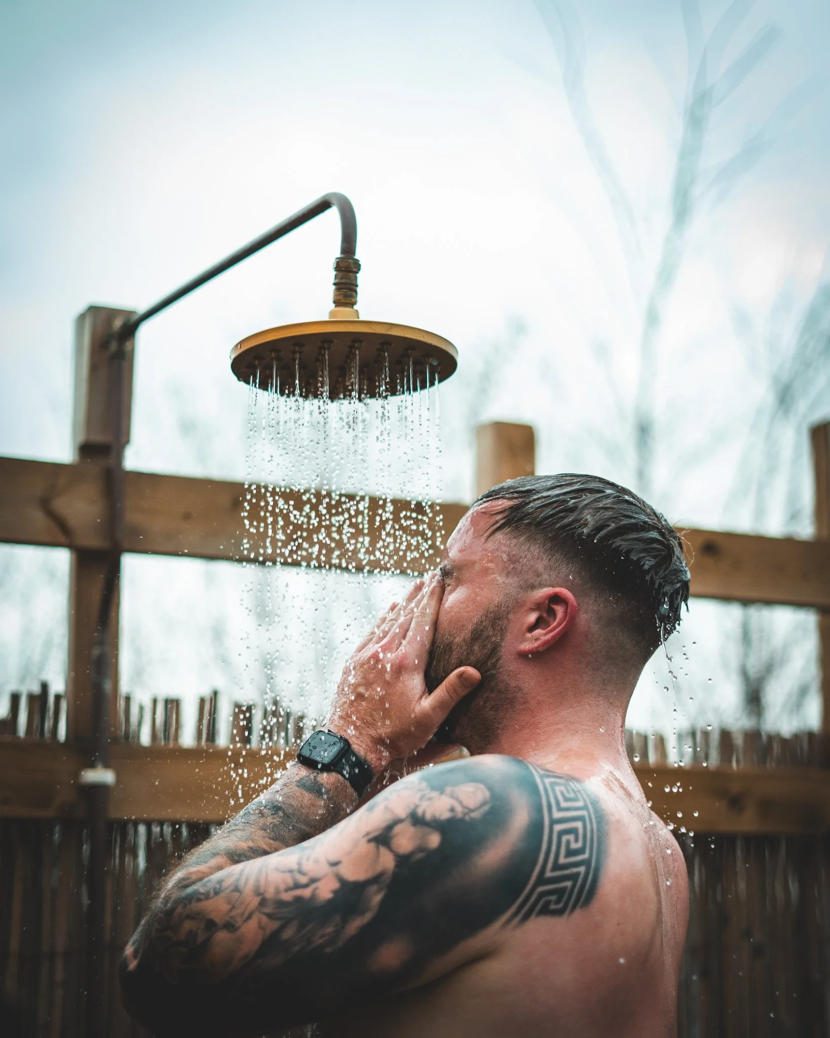 At Unfold Sauna Club Man taking an outdoor shower with water falling from a showerhead, tattoos on his arm, wearing a wristwatch, and a wooden fence in the background.