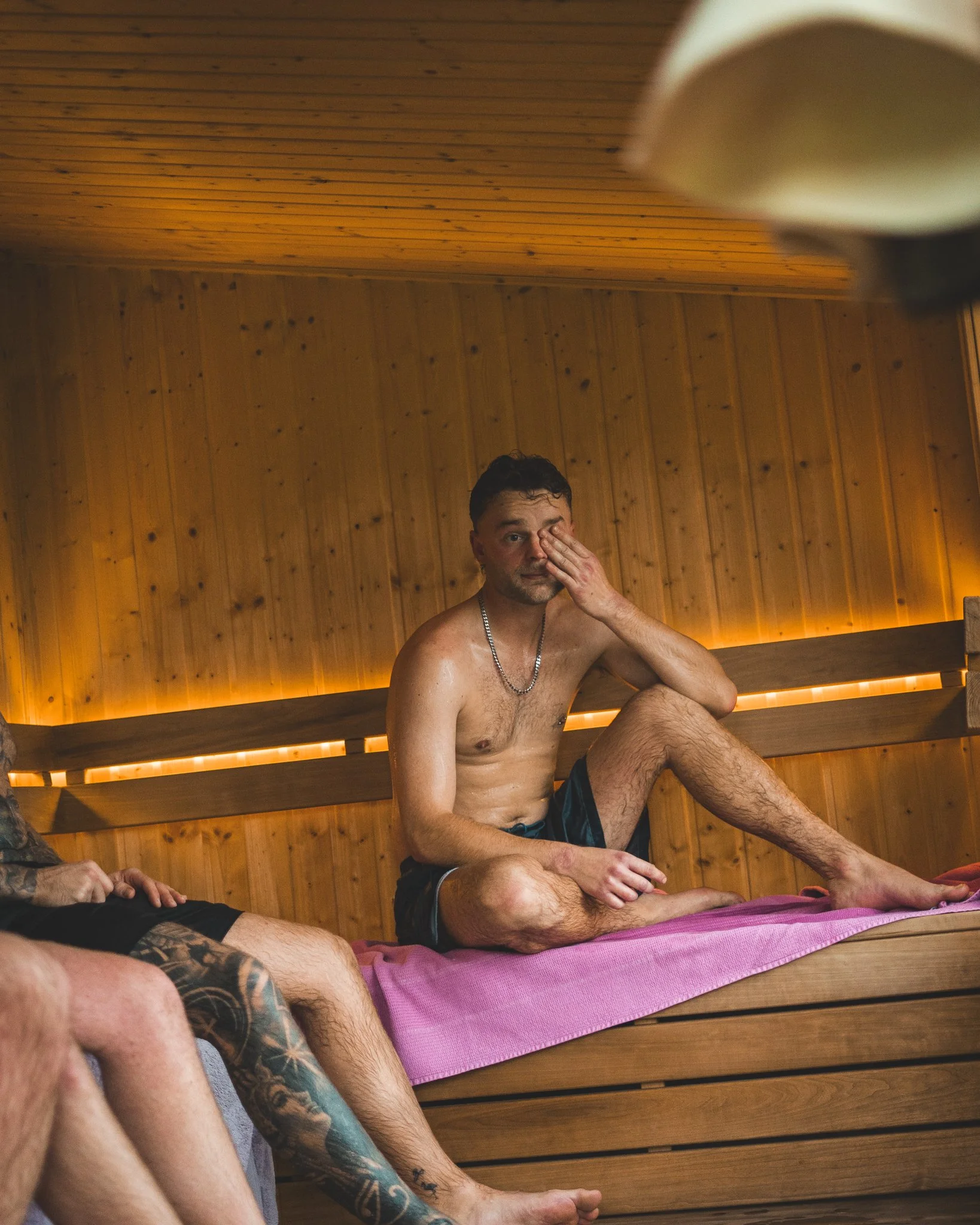 At Unfold Sauna Club A shirtless man sitting inside a sauna with wooden walls and benches, touching his face with one hand, surrounded by other partially visible men with tattoos.