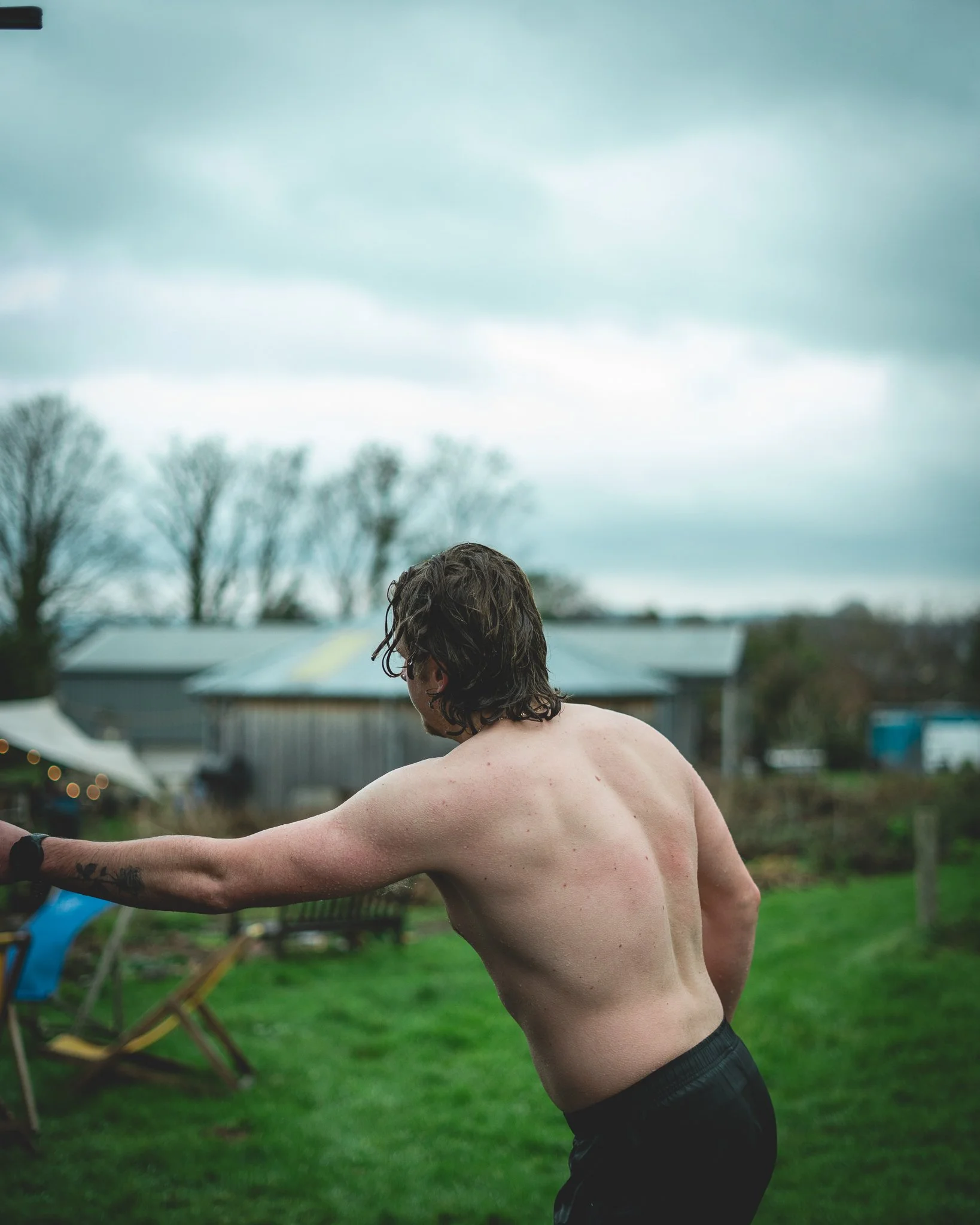 At Unfold Sauna Club A shirtless man with wet hair stretching his arm outdoors on a cloudy day, with a grassy yard, trees, and a barn in the background.
