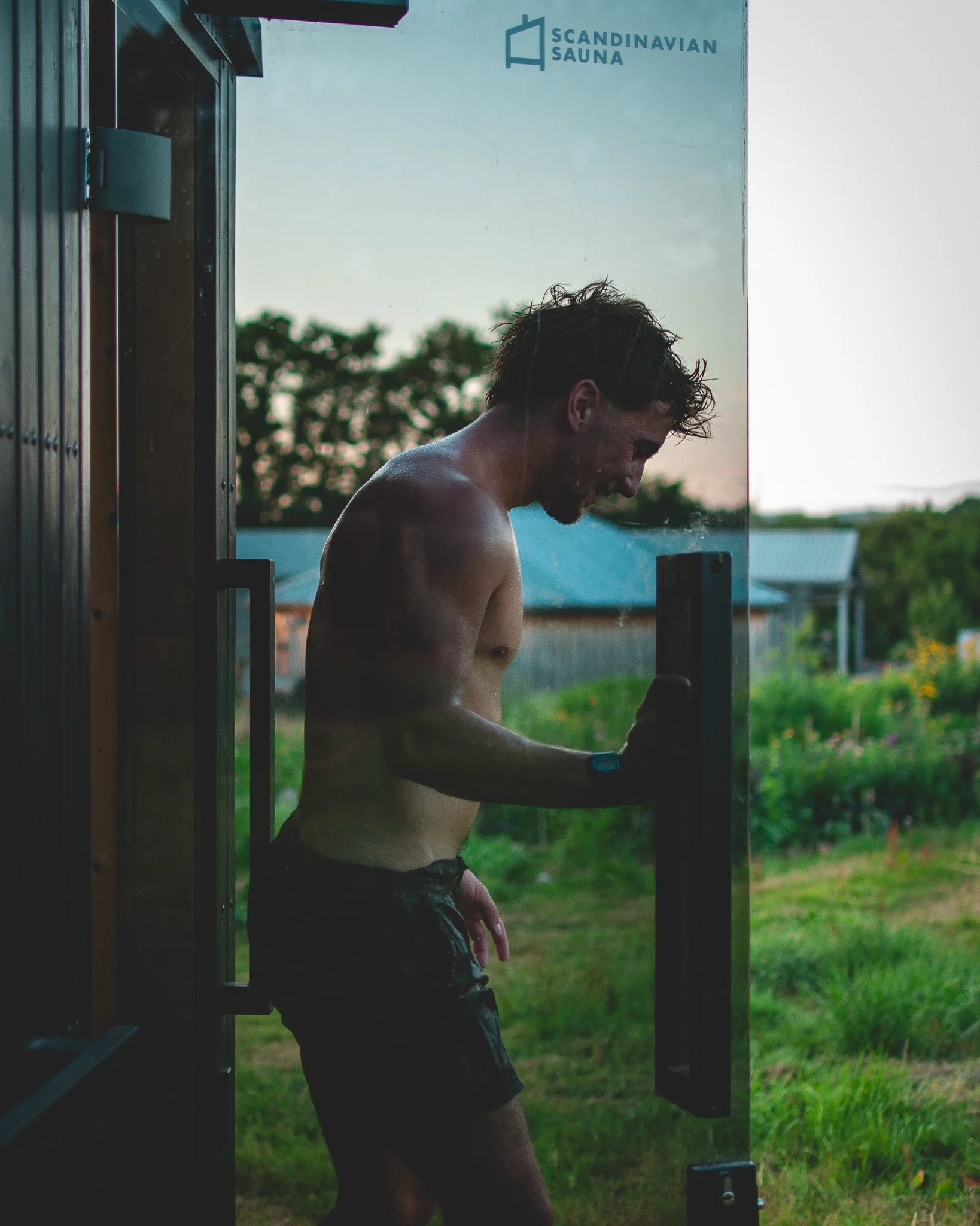 At Unfold Sauna Club A shirtless man with dark, wet hair opening a door to a Scandinavian sauna, with a grassy outdoor area and trees in the background during sunset.