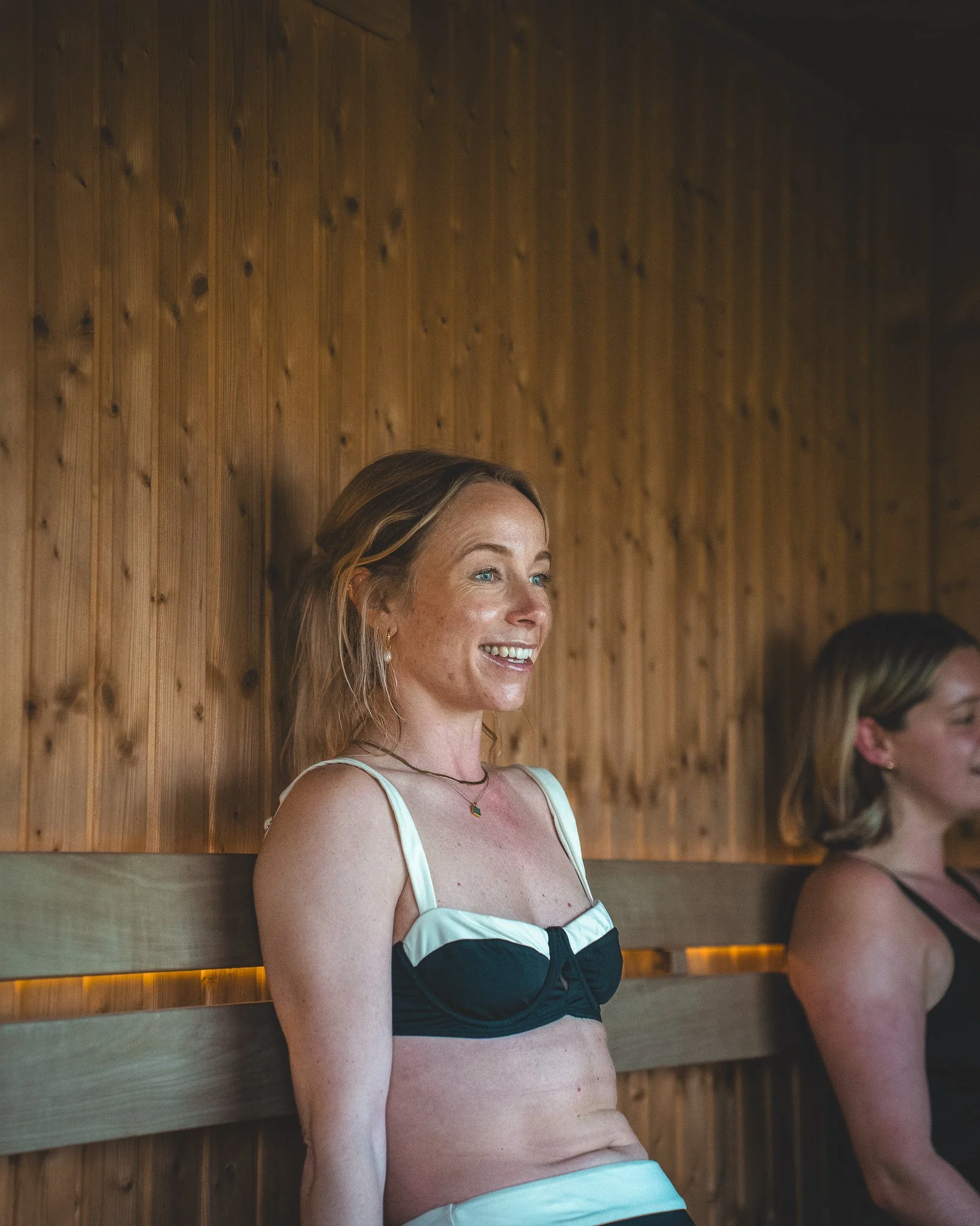 At Unfold Sauna Club A woman wearing a black and white bikini top, smiling and leaning against a wooden wall in a sauna.