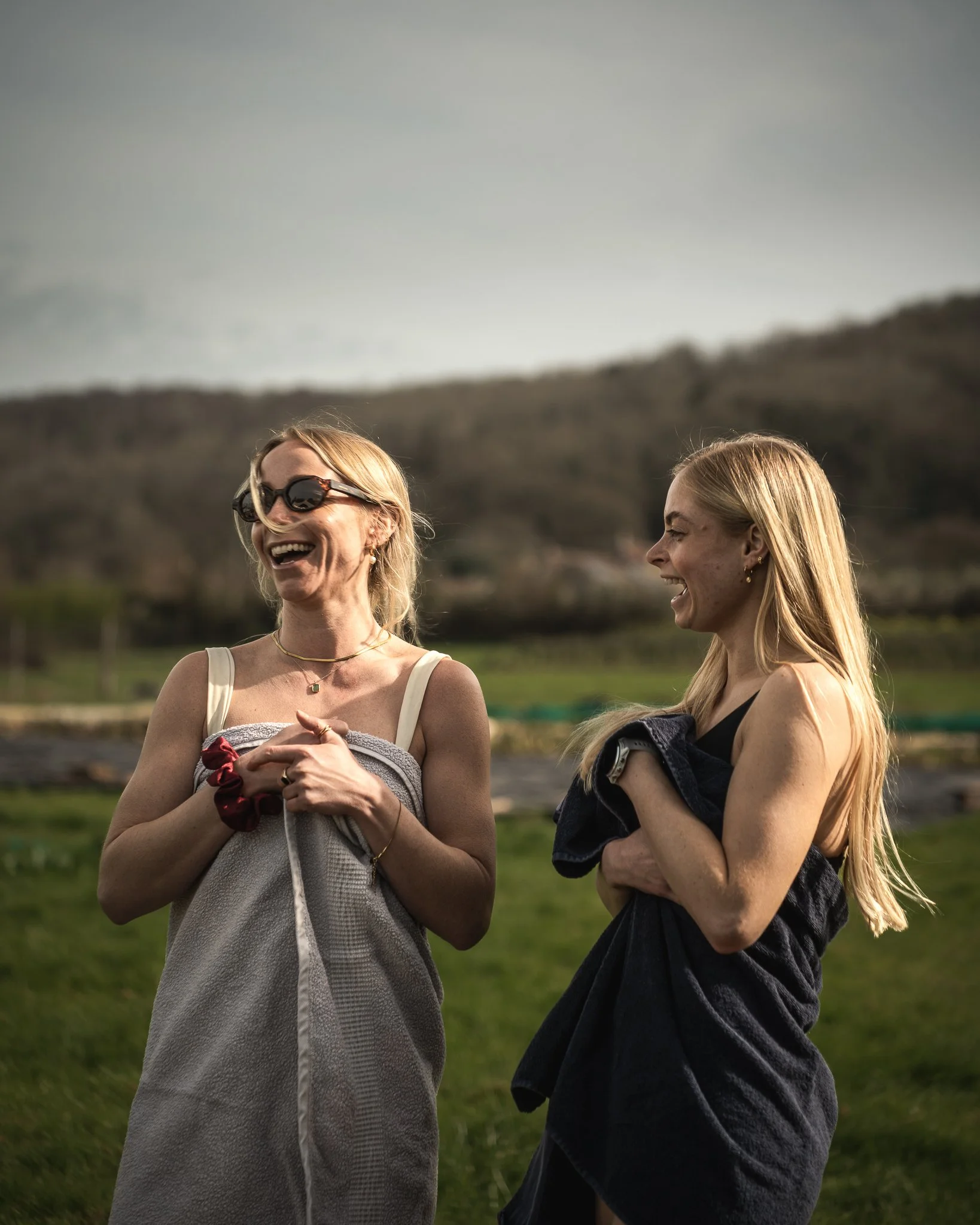 At Unfold Sauna Club Two women outdoors, wrapped in towels, laughing and enjoying each other's company, with a grassy field and hills in the background.