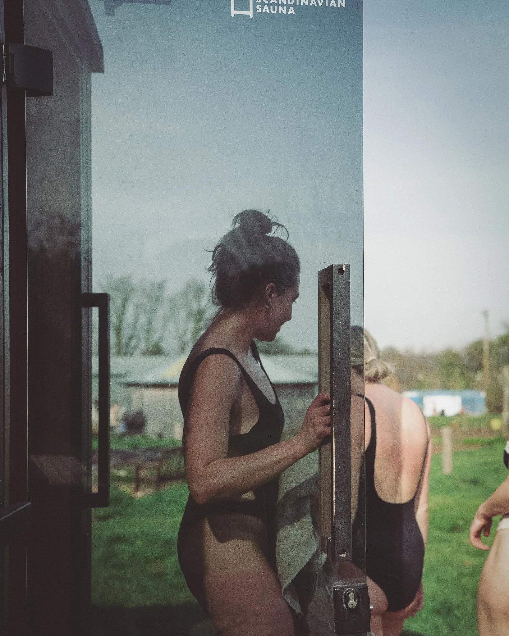 At Unfold Sauna Club A woman in a black swimsuit standing outside near a sauna door, holding a towel and looking down, with two other women in swimsuits nearby, in a grassy outdoor setting.