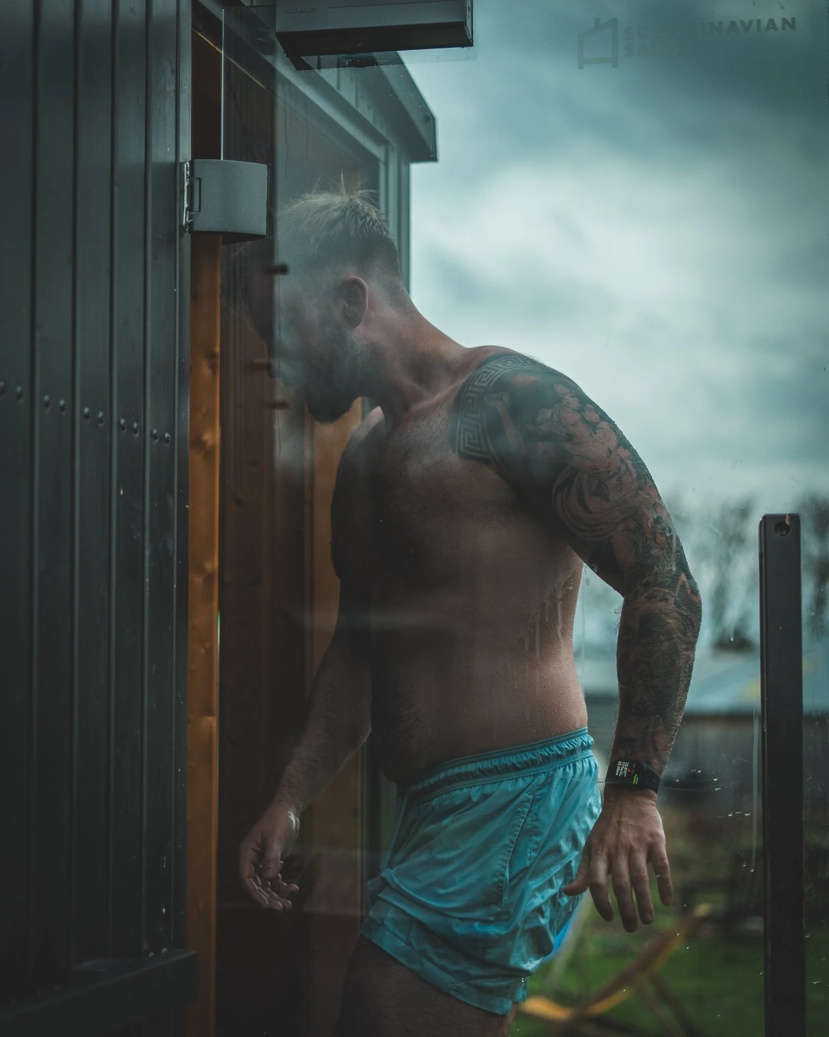 At Unfold Sauna Club A muscular man with tattoos standing shirtless outside a sauna with a glass door, looking inside.