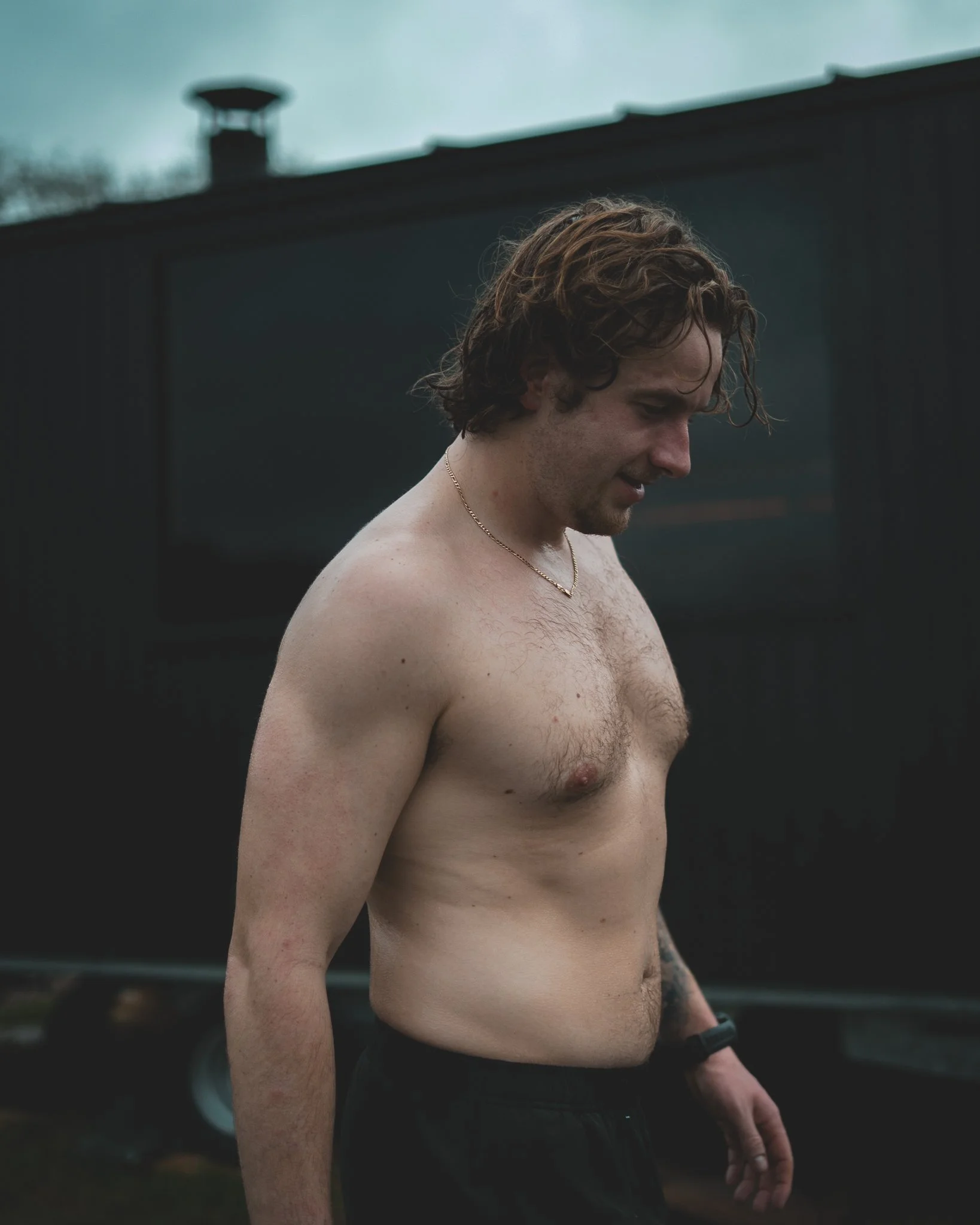 At Unfold Sauna Club A shirtless man with curly brown hair looking downward outside next to a dark building.