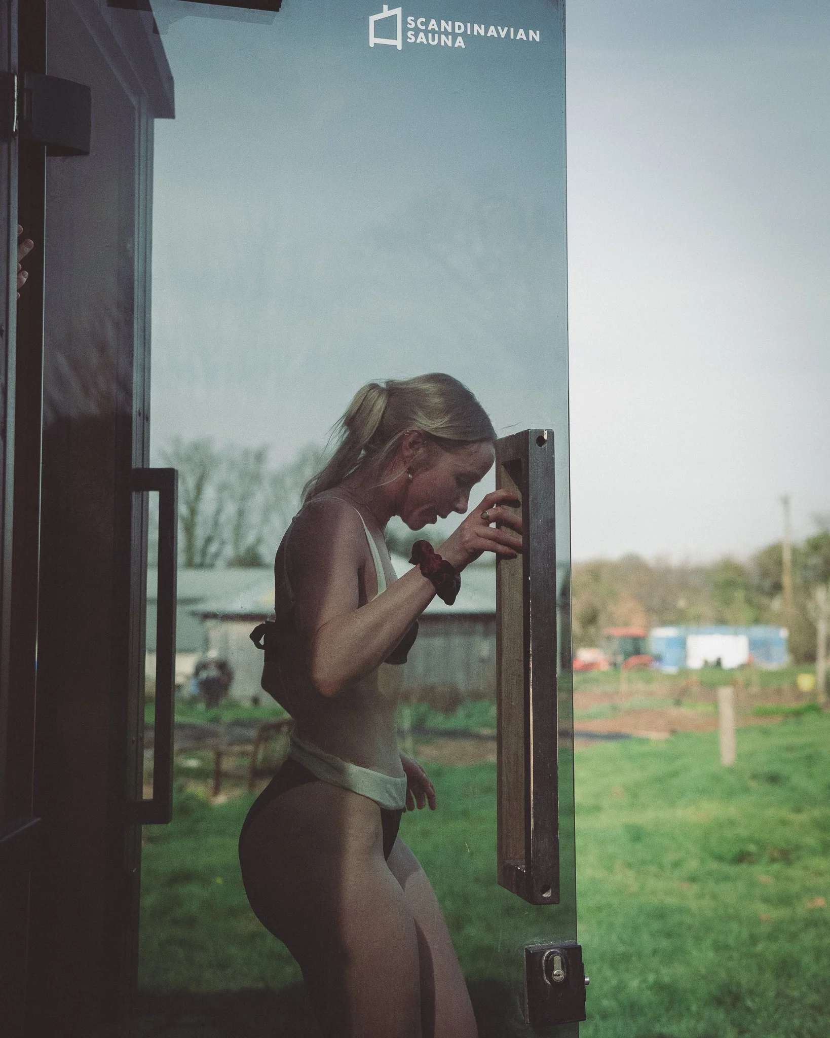 At Unfold Sauna Club A woman standing inside a sauna door, looking down, with a green outdoor landscape in the background.