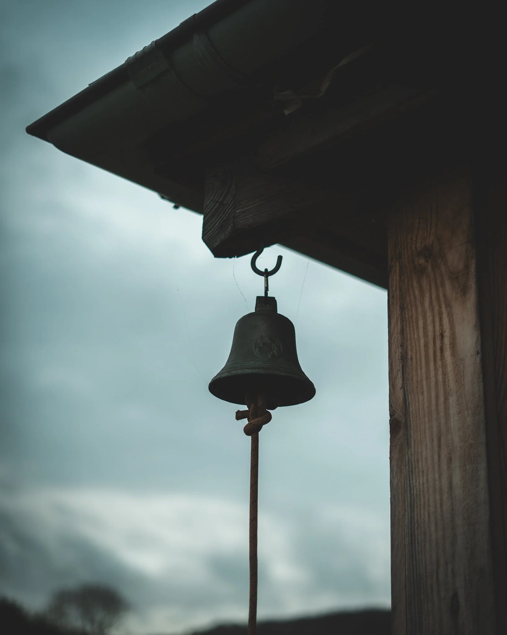 At Unfold Sauna Club A small bell hanging from a hook under the eave of a house, with a rope looped through its clapper, against a cloudy sky background.