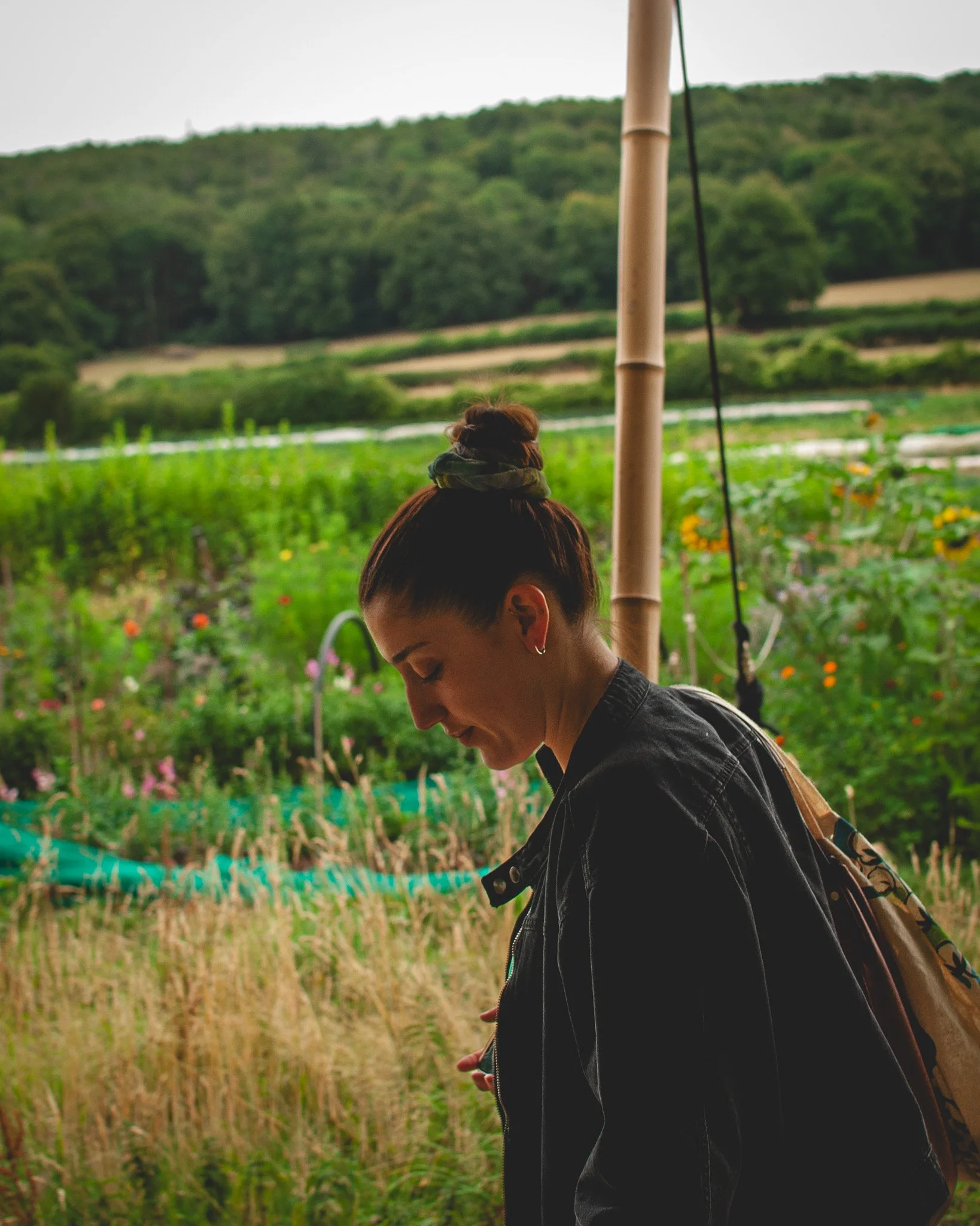 At Unfold Sauna Club Woman with hair tied back, wearing a black jacket and patterned bag, looking down in a garden with flowers and greenery, with hills in background.
