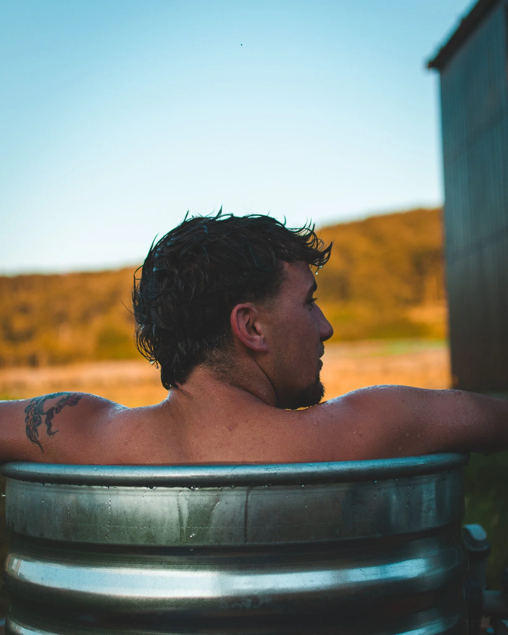 At Unfold Sauna Club A man with dark, curly hair and a tattoo on his arm is relaxing in a metal hot tub outdoors, with a backdrop of hills and a clear sky.