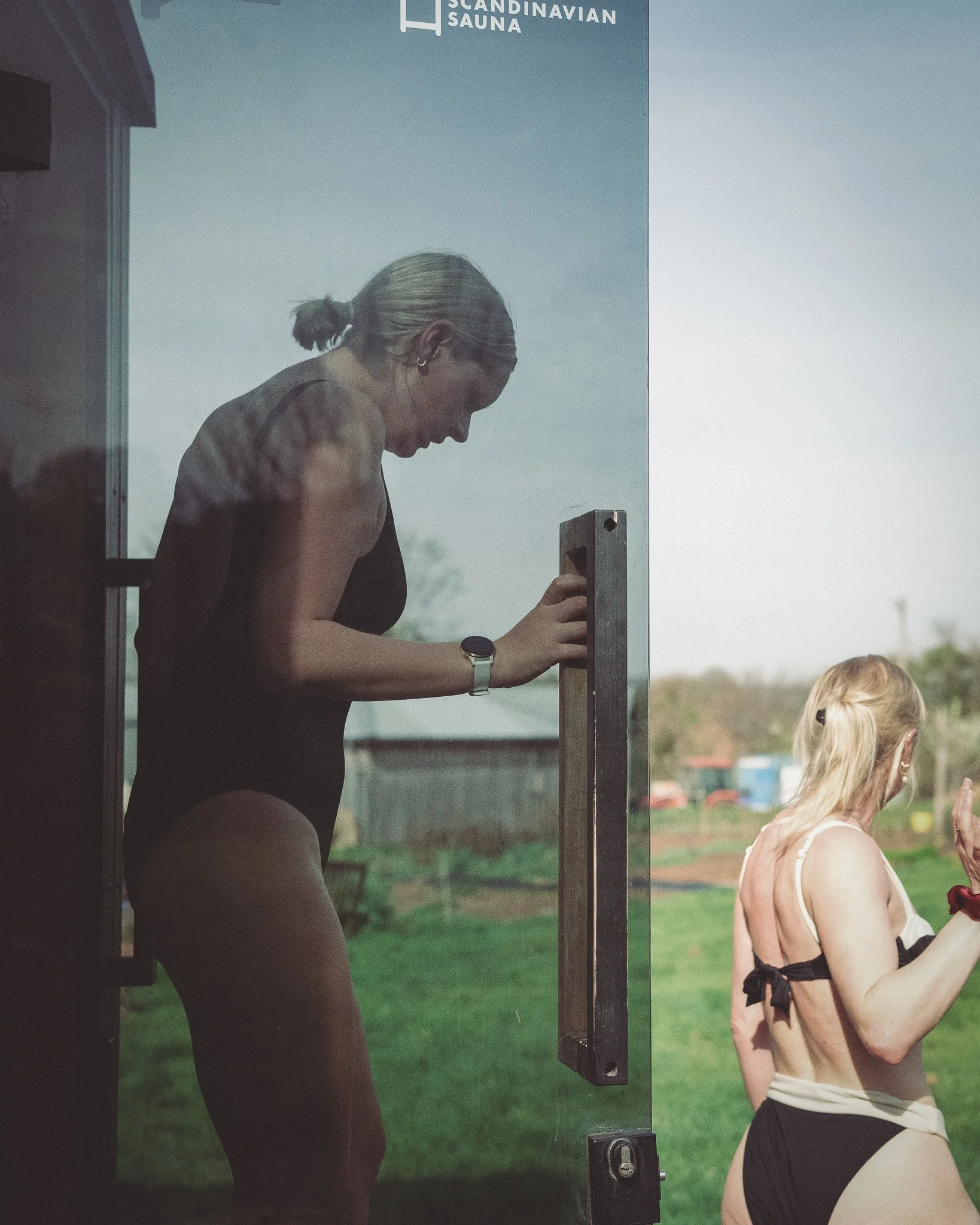 At Unfold Sauna Club A woman in a black swimsuit is opening a glass door, and a woman in a black and white swimsuit is facing away outside on a grassy field.
