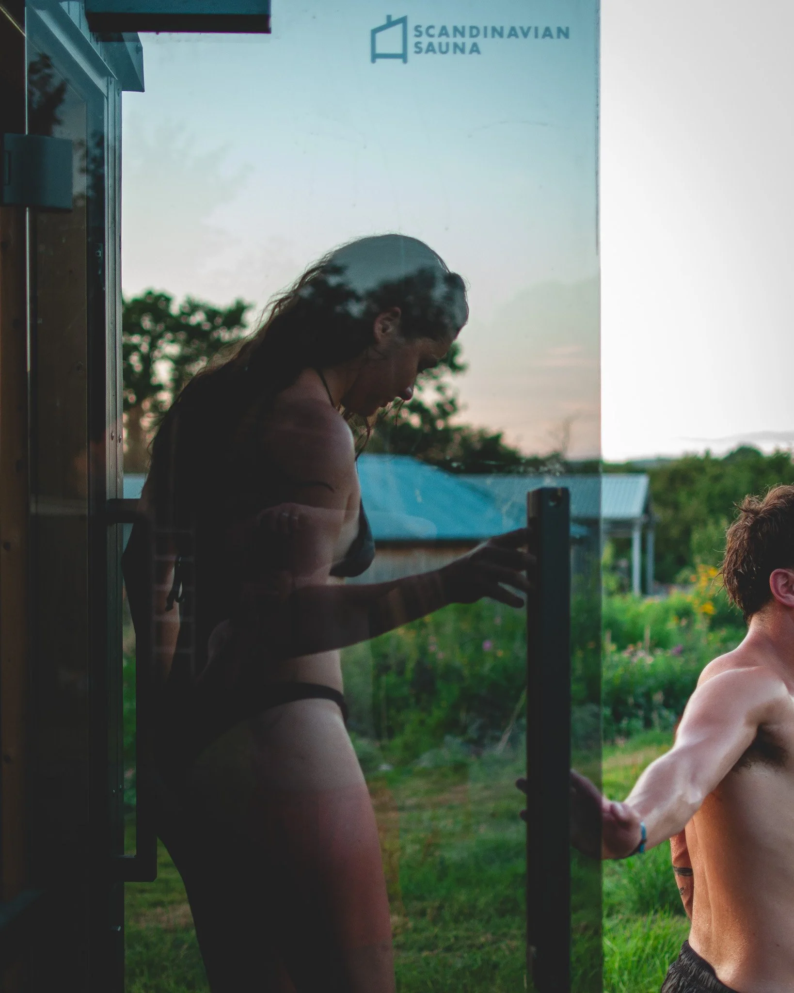 A woman is seen through a glass door, with her silhouette visible against a scenic outdoor background. She appears to be taking a photo or looking at something on her phone. The outdoor scene features greenery and a small building, with a sign that r