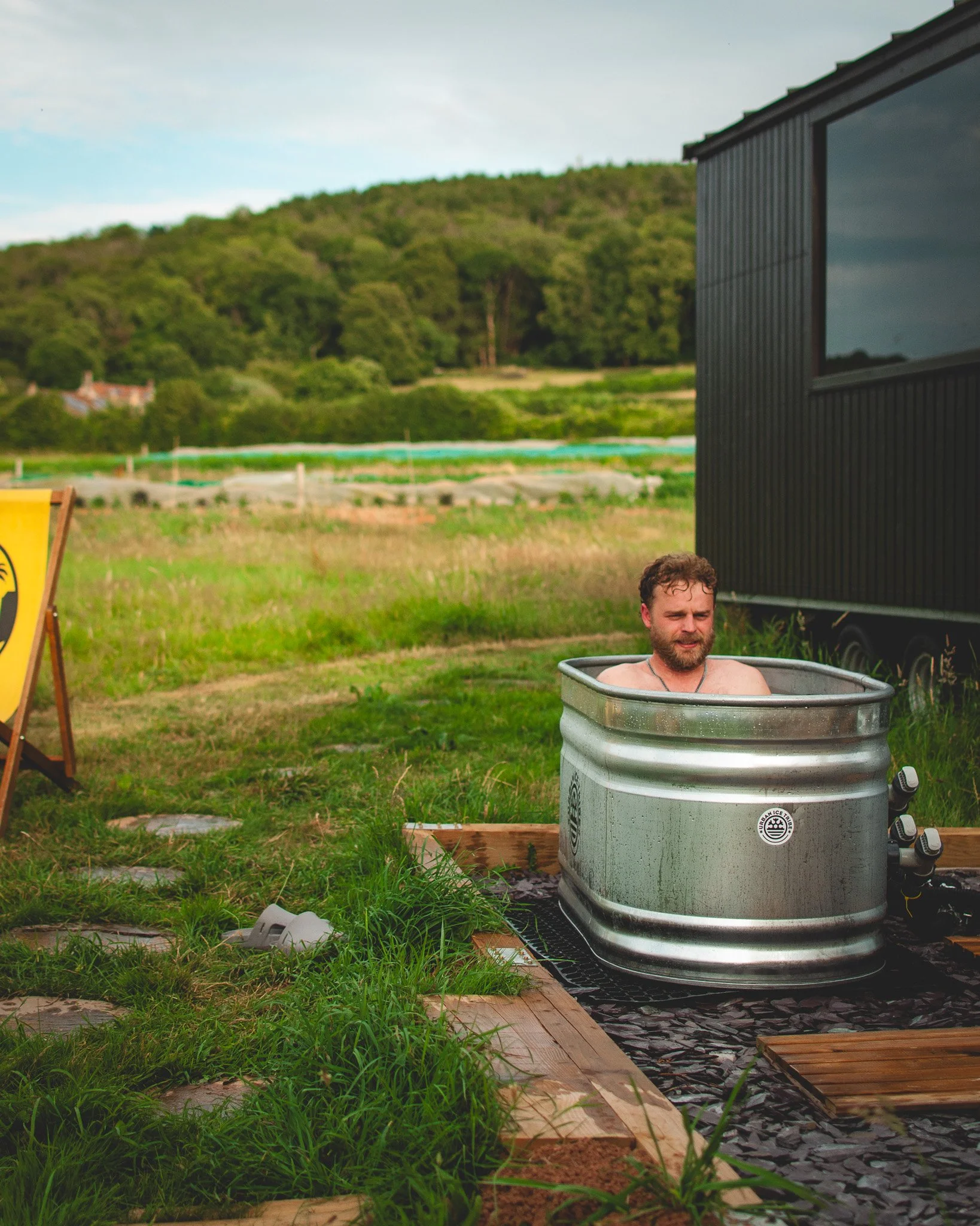 At Unfold Sauna Club A man sitting in a metal bathtub outdoors on a grassy area during the daytime, surrounded by a black wooden building and mountains in the background.
