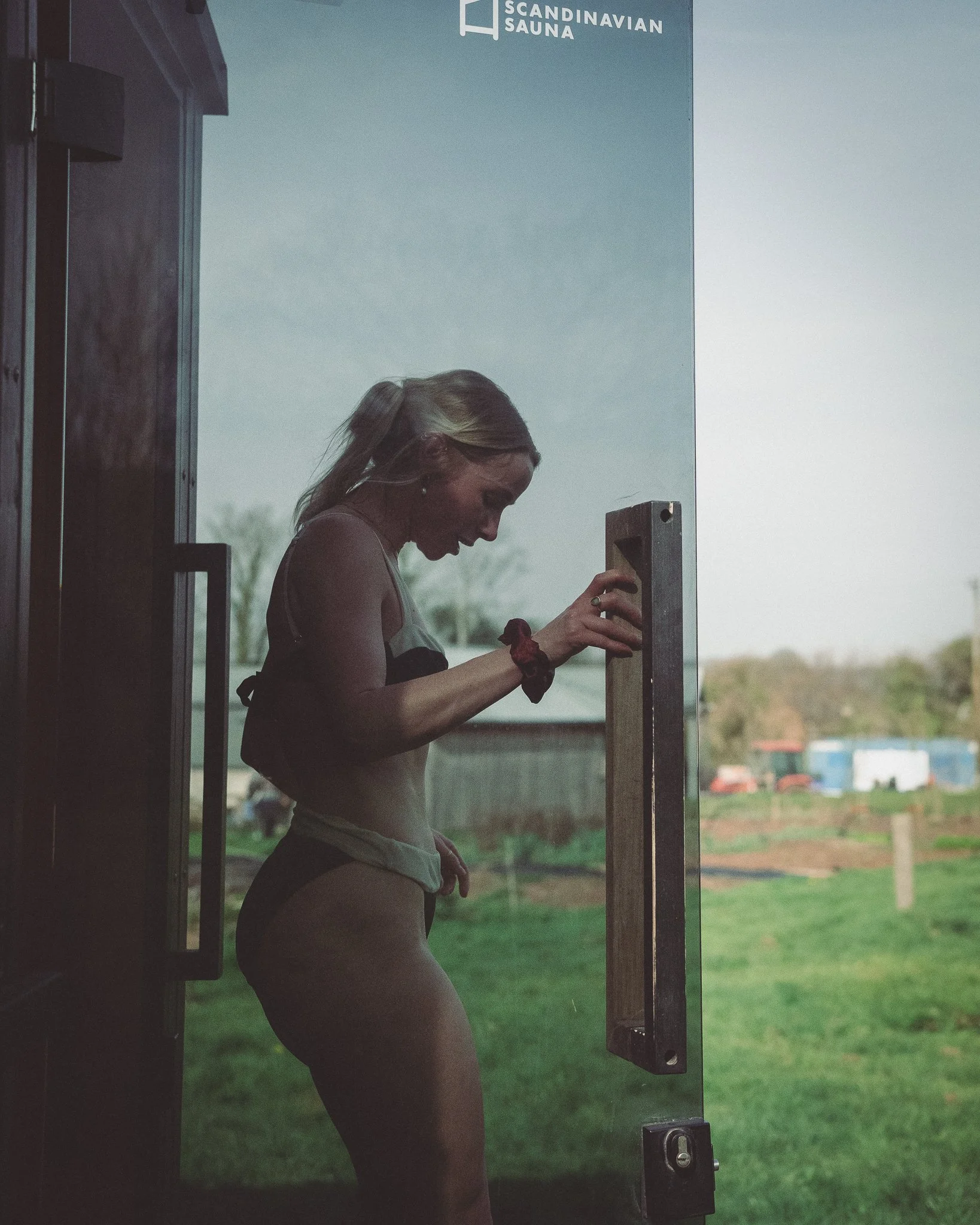 At Unfold Sauna Club A woman standing at the door of a Scandinavian Sauna, looking down, with her hand on the door handle inside the sauna.