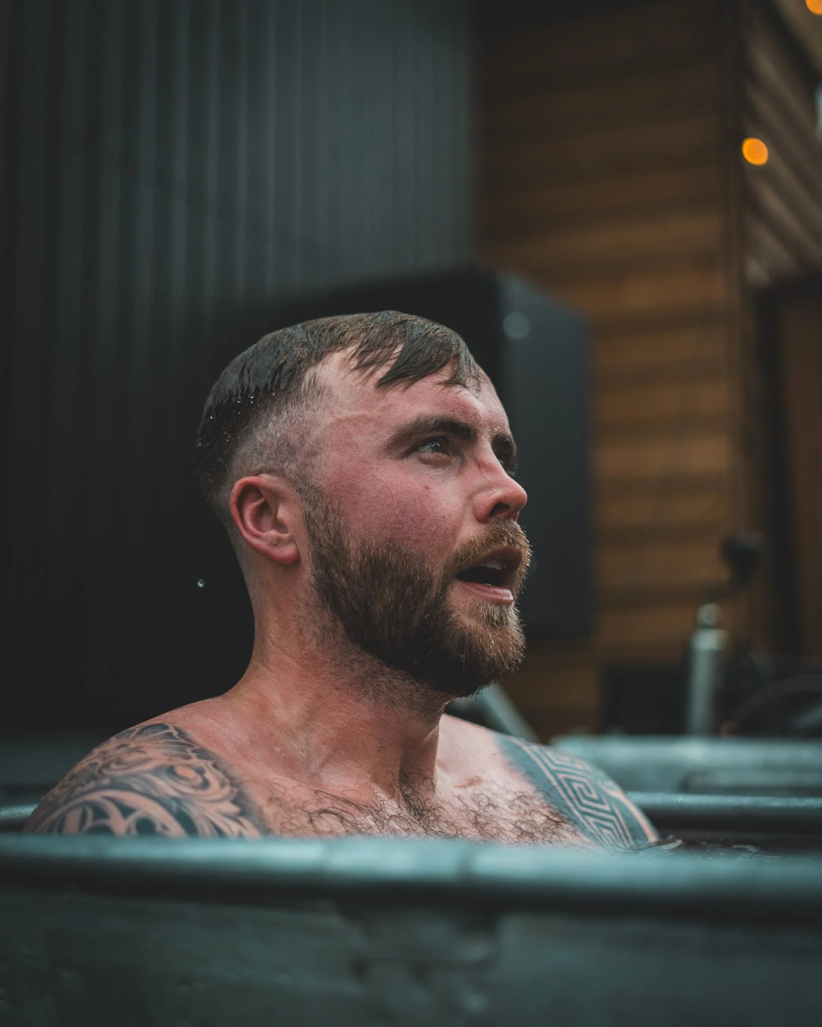 At Unfold Sauna Club A man with a beard and tattoos on his shoulder is soaking in a bathtub, looking upwards with a thoughtful expression, in a bathroom setting.